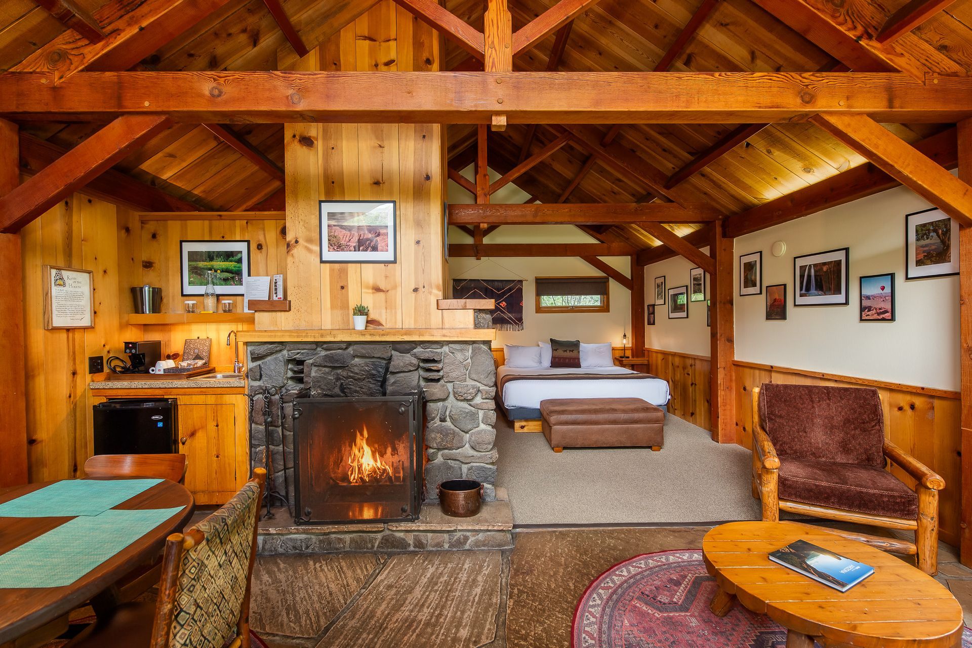 Cozy cabin interior: stone fireplace with fire, bed, sitting area, kitchenette, wooden beams, and warm lighting.