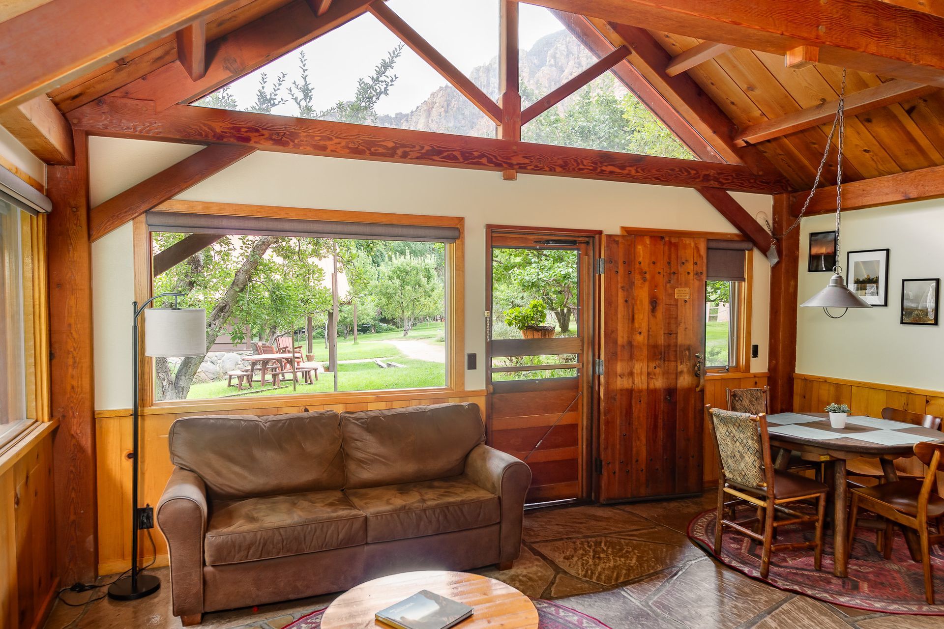 Cozy cabin interior with exposed wood beams, sofa, dining table, and large windows overlooking a green outdoor area.