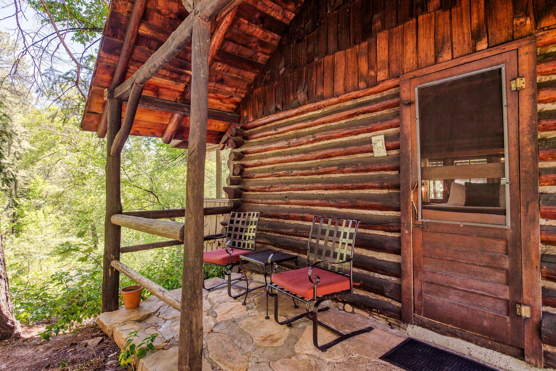 Log cabin exterior with porch, two chairs, and door. Wood and stone in forest setting.