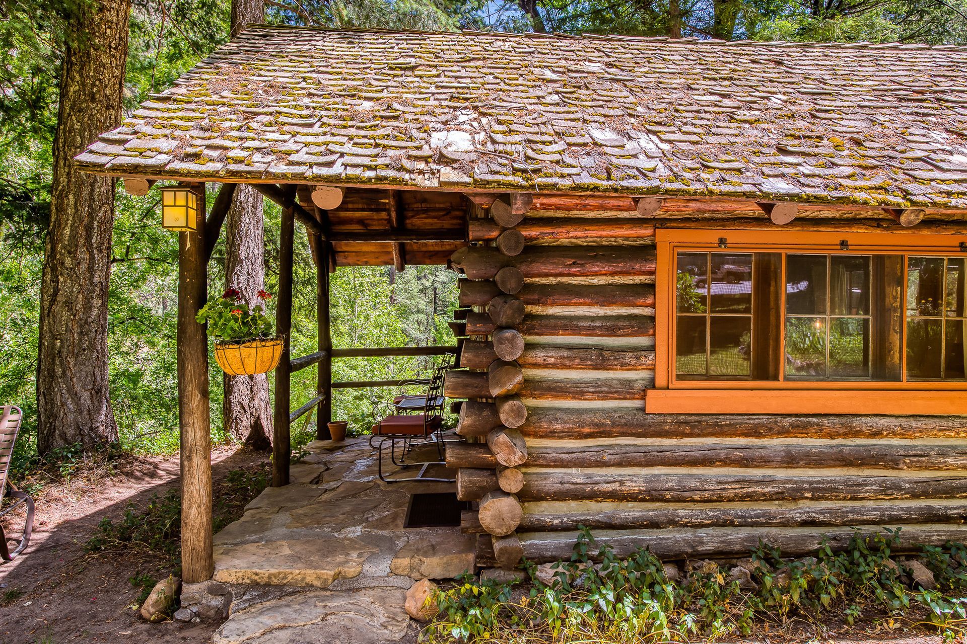 Log cabin with a moss-covered roof, porch, and orange-framed window, nestled in a wooded area.