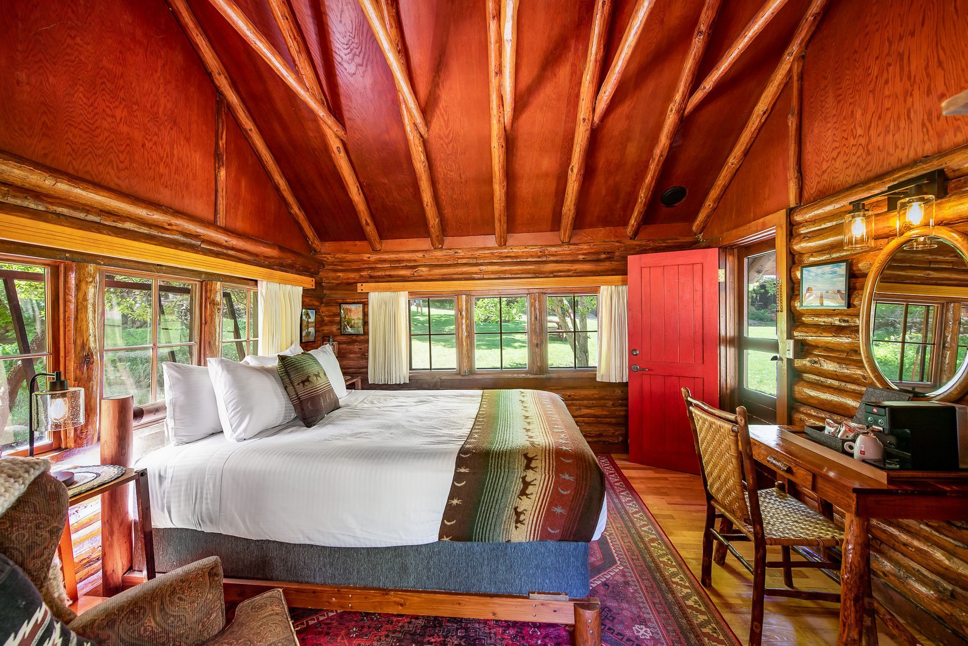 Cozy cabin bedroom with a bed, desk, and red door. Log walls and ceiling, windows with a view.