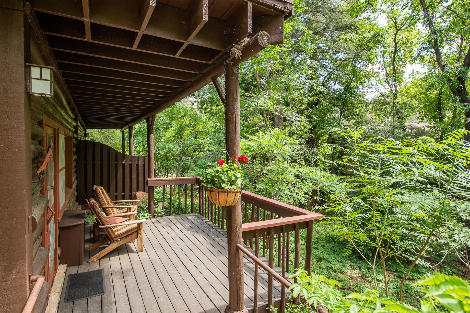 Wooden porch with two chairs, railing, and hanging flower basket, overlooking lush green trees.