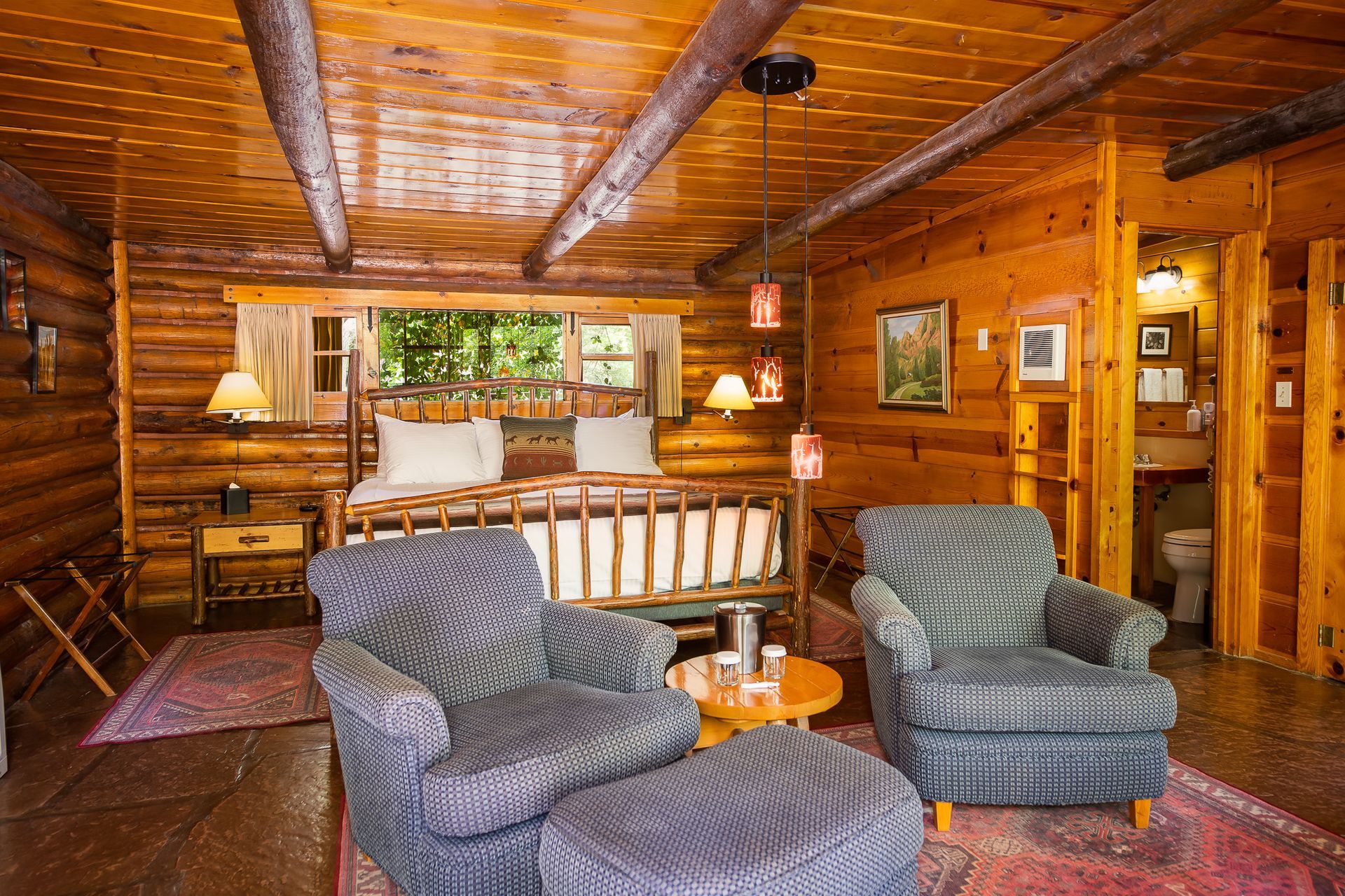 Rustic log cabin bedroom with two armchairs, bed, and open doorway to a bathroom.