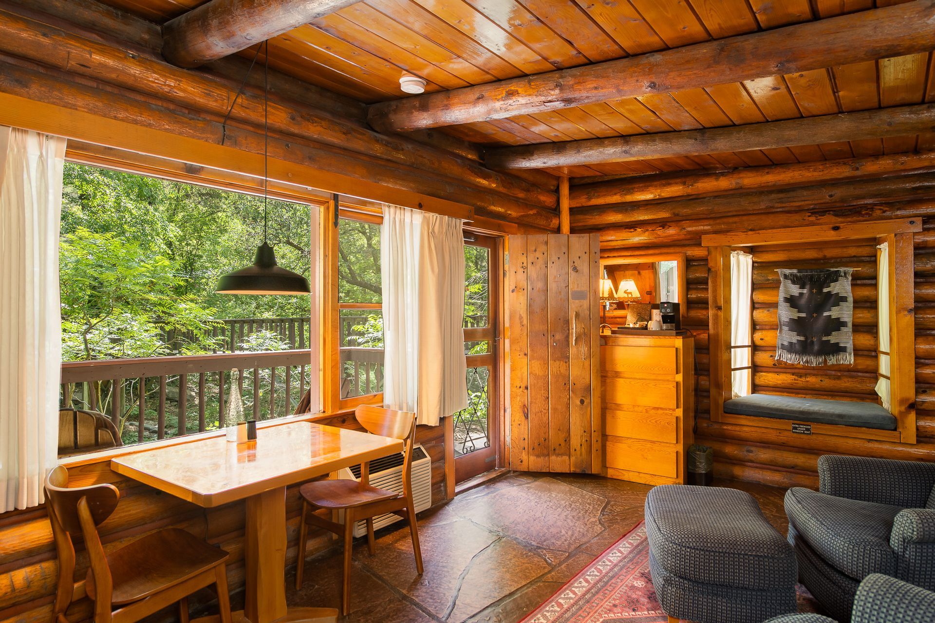 Cozy cabin interior: wooden walls, window overlooking trees, table with chairs, and a small couch.