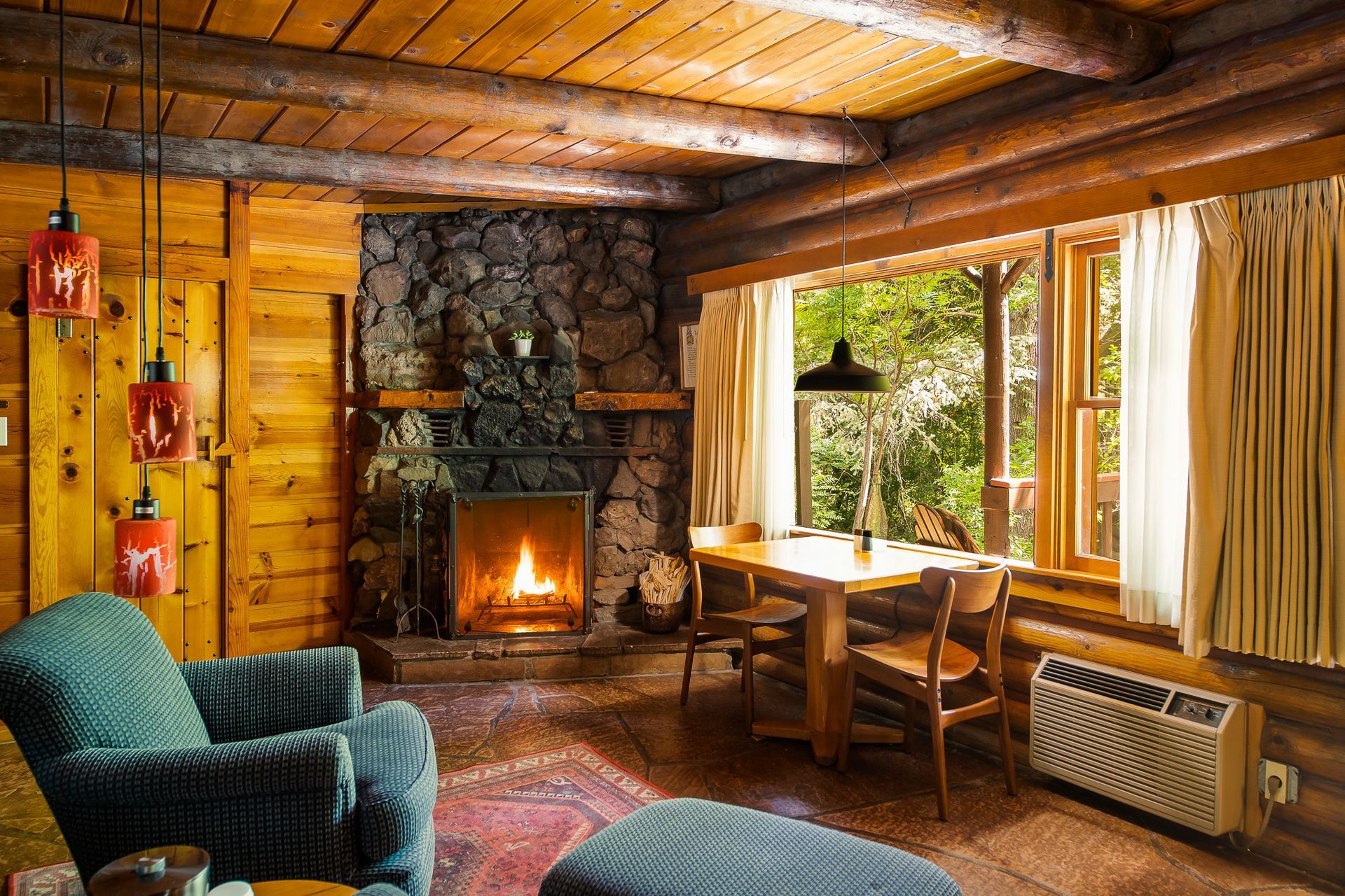 Cozy cabin interior with stone fireplace, wooden beams, table by window, and teal armchair.