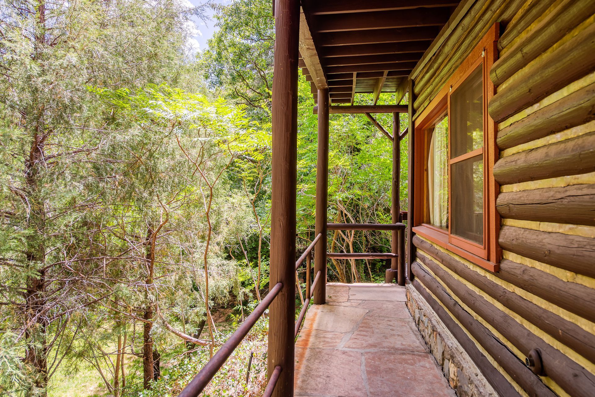 Log cabin porch with railing and window overlooking lush green trees.