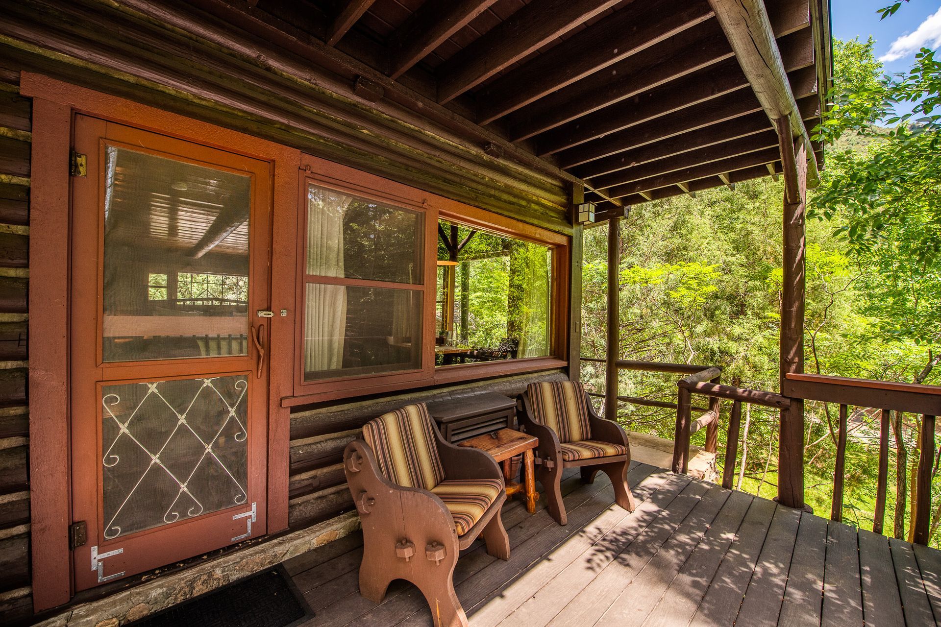 Wooden cabin porch with two chairs and a small table, overlooking lush green trees.