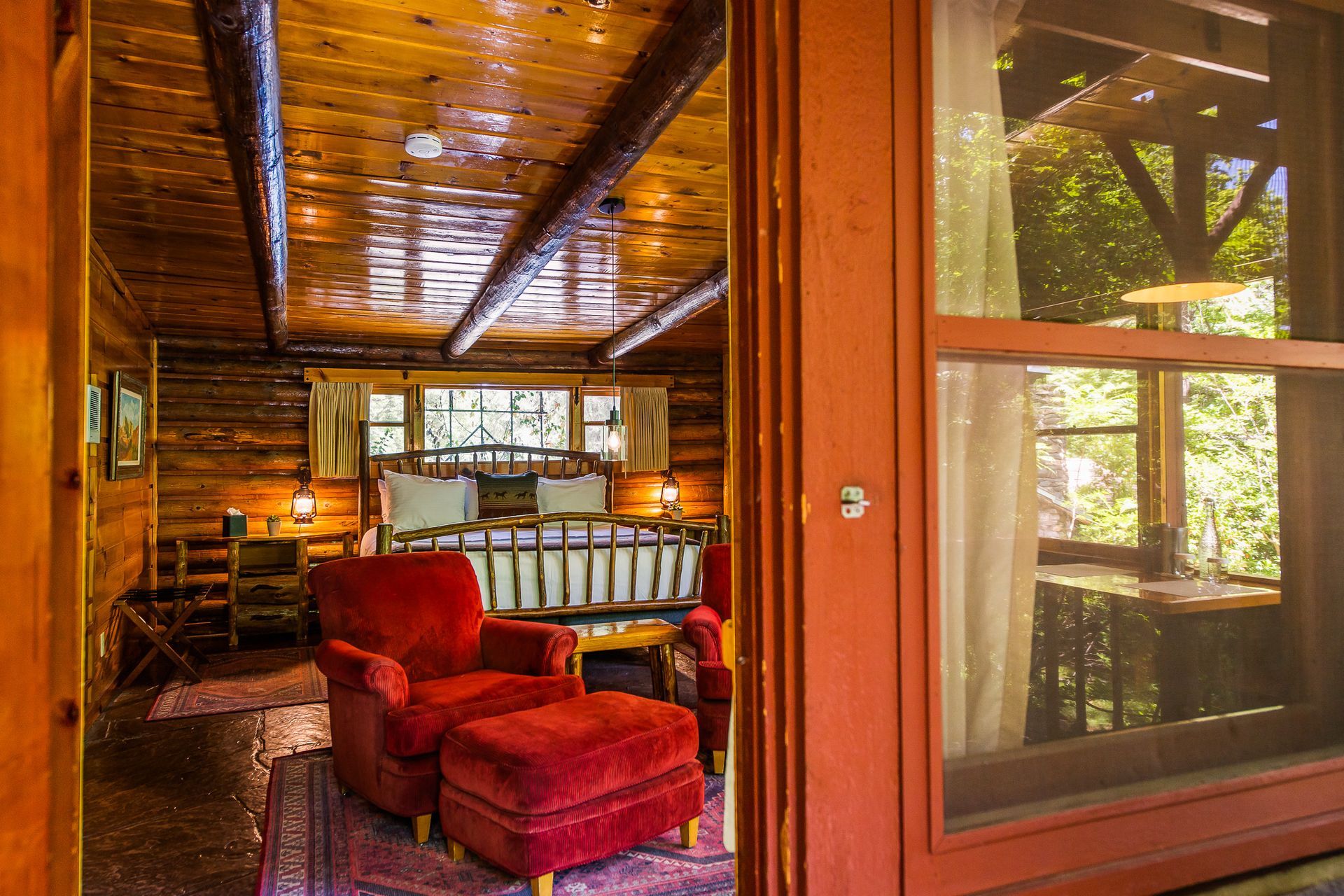 Cozy cabin bedroom with a red armchair and ottoman, a double bed, and a window overlooking trees.