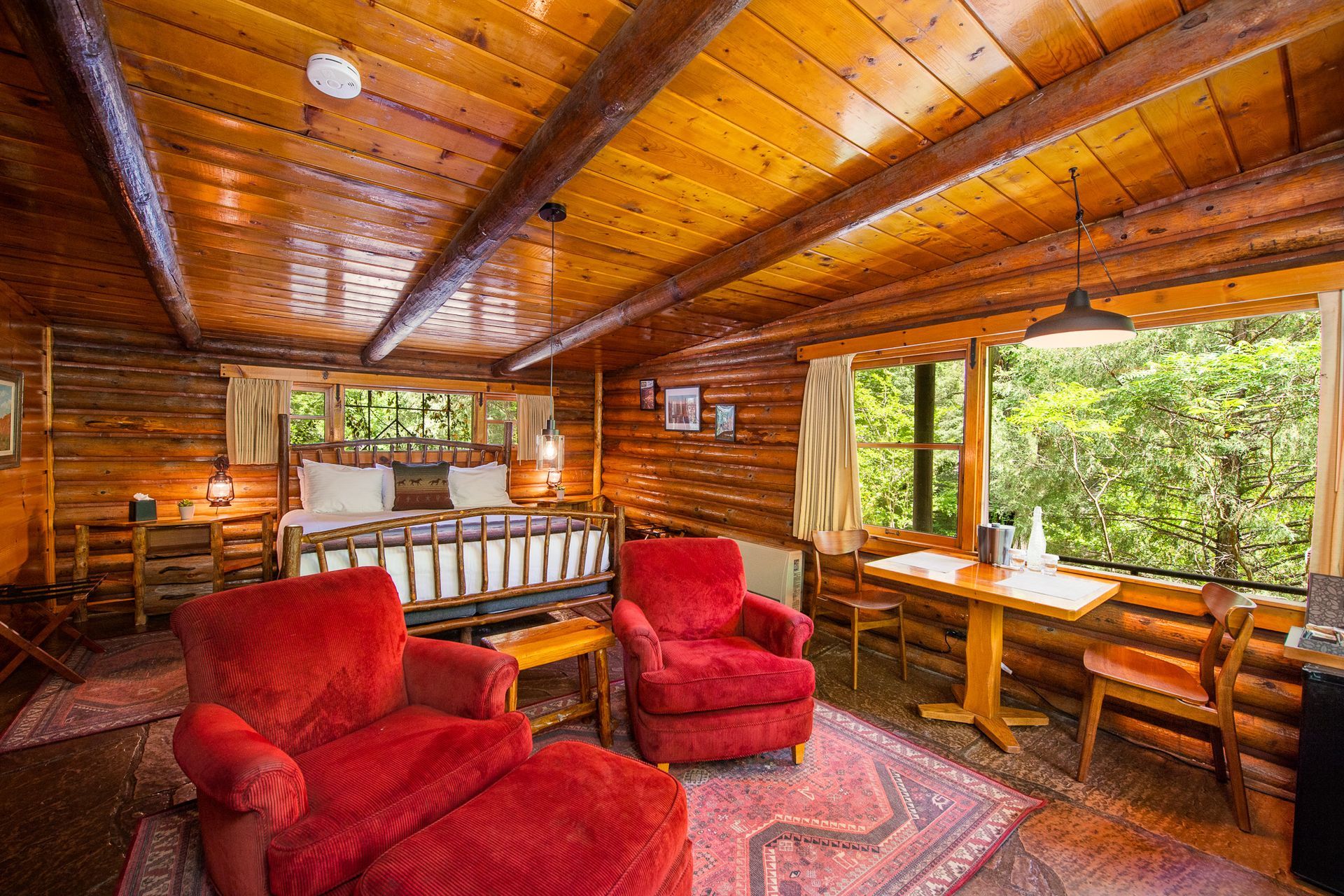Rustic cabin interior with a bed, red armchairs, and a small table near a window.
