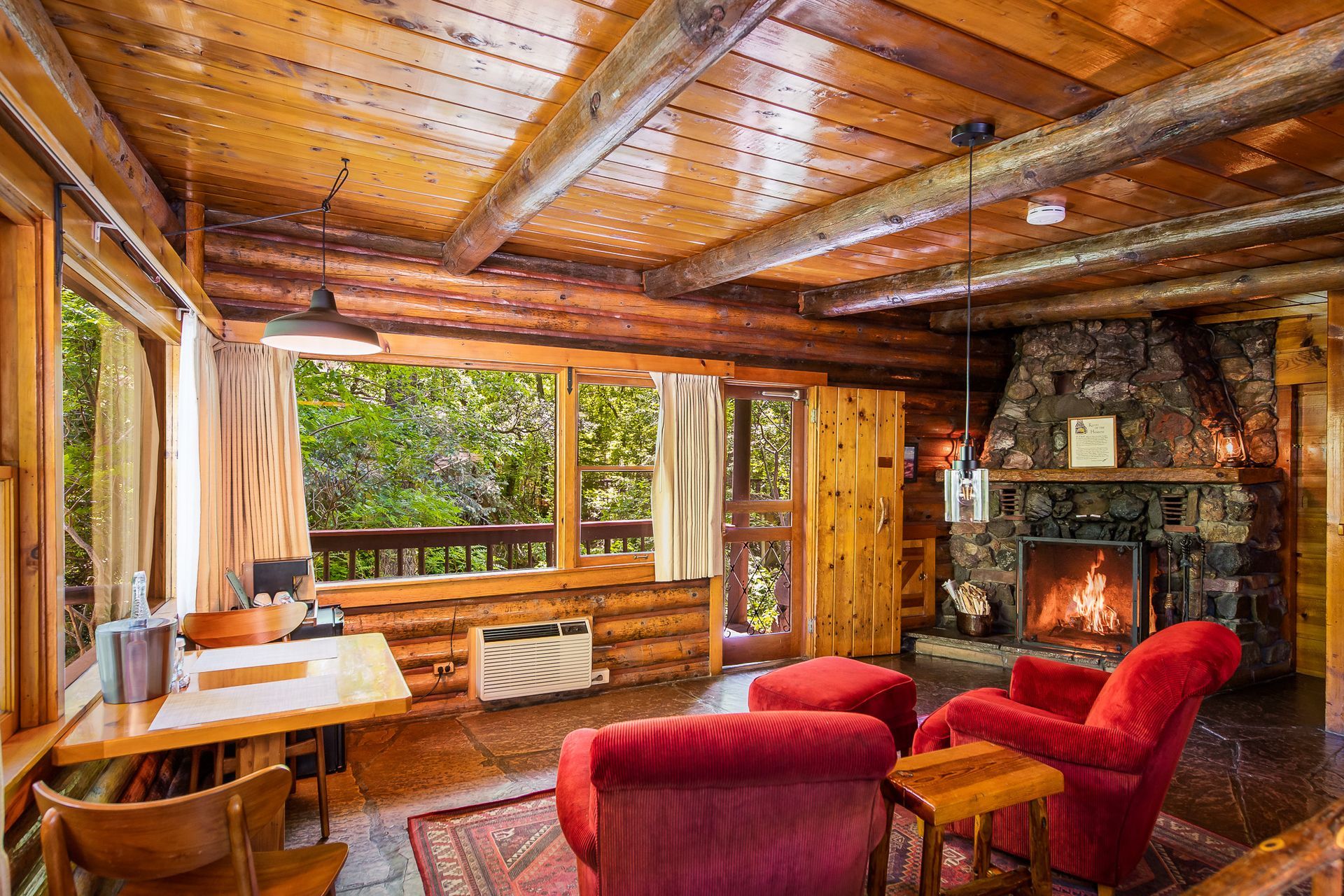 Cozy cabin interior: log walls, stone fireplace, red armchairs, and a view of trees through the windows.