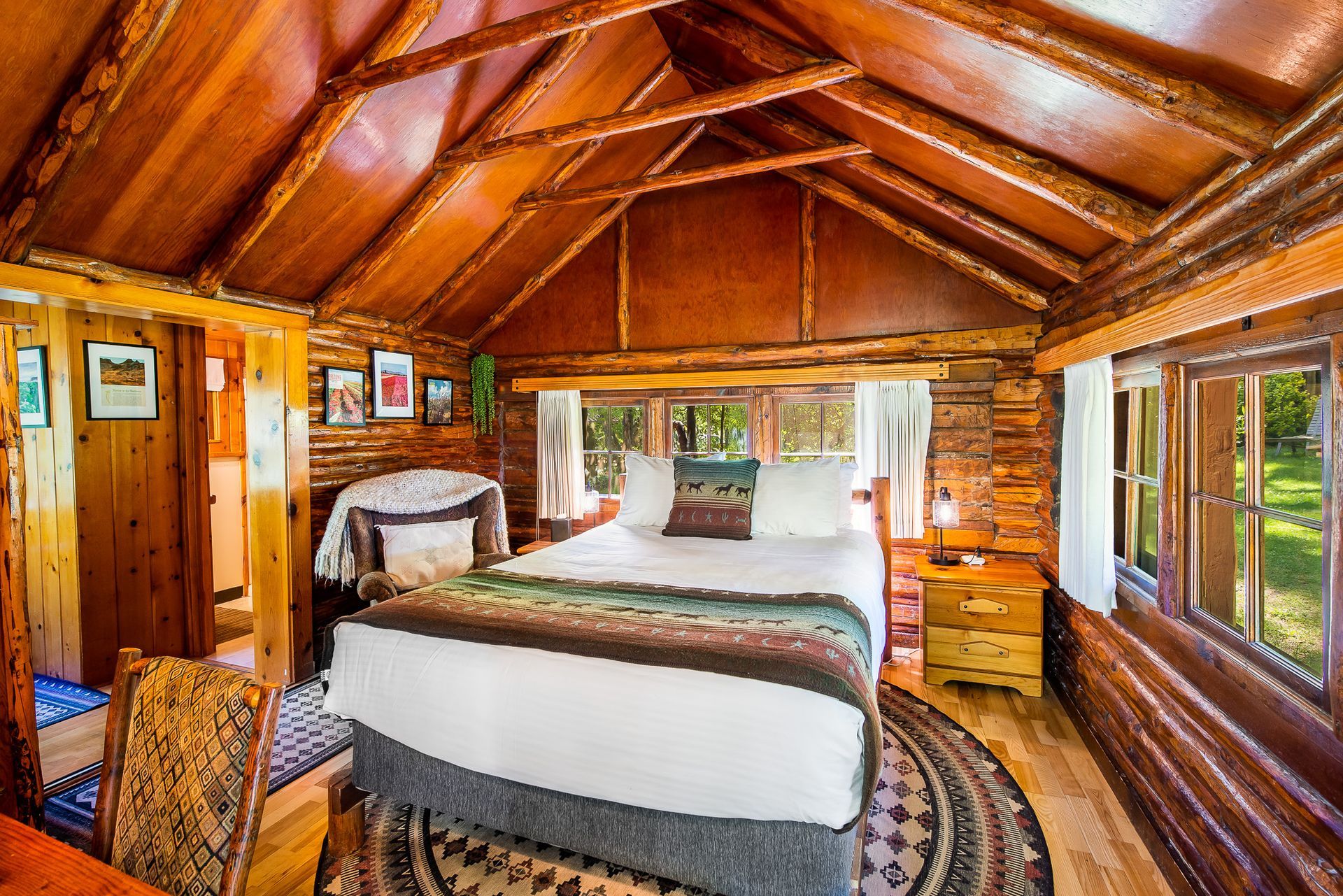Cozy cabin bedroom with a log ceiling and walls, bed, chair, and windows looking out at nature.