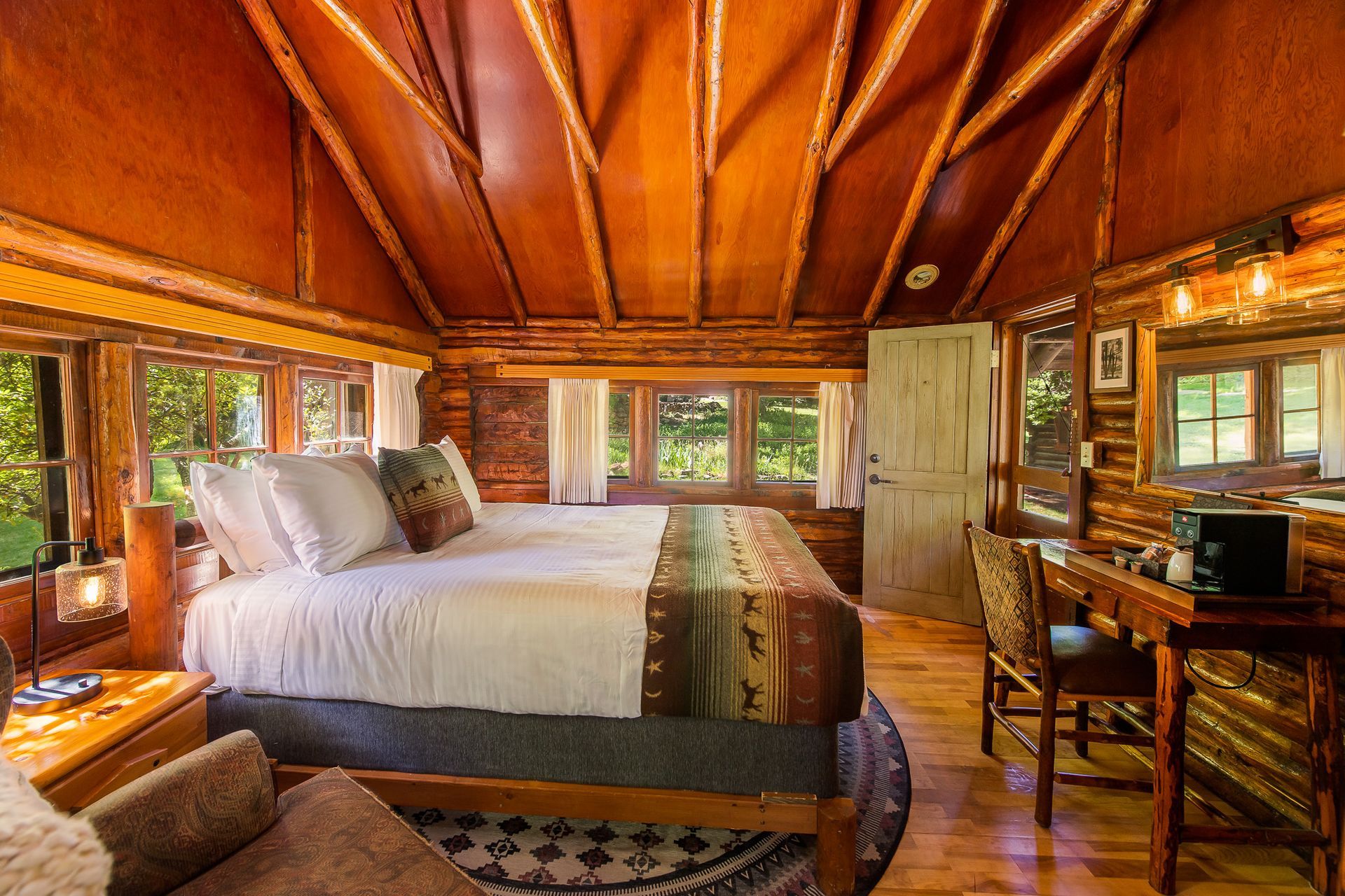 Cozy log cabin bedroom with a bed, desk, and multiple windows overlooking trees.