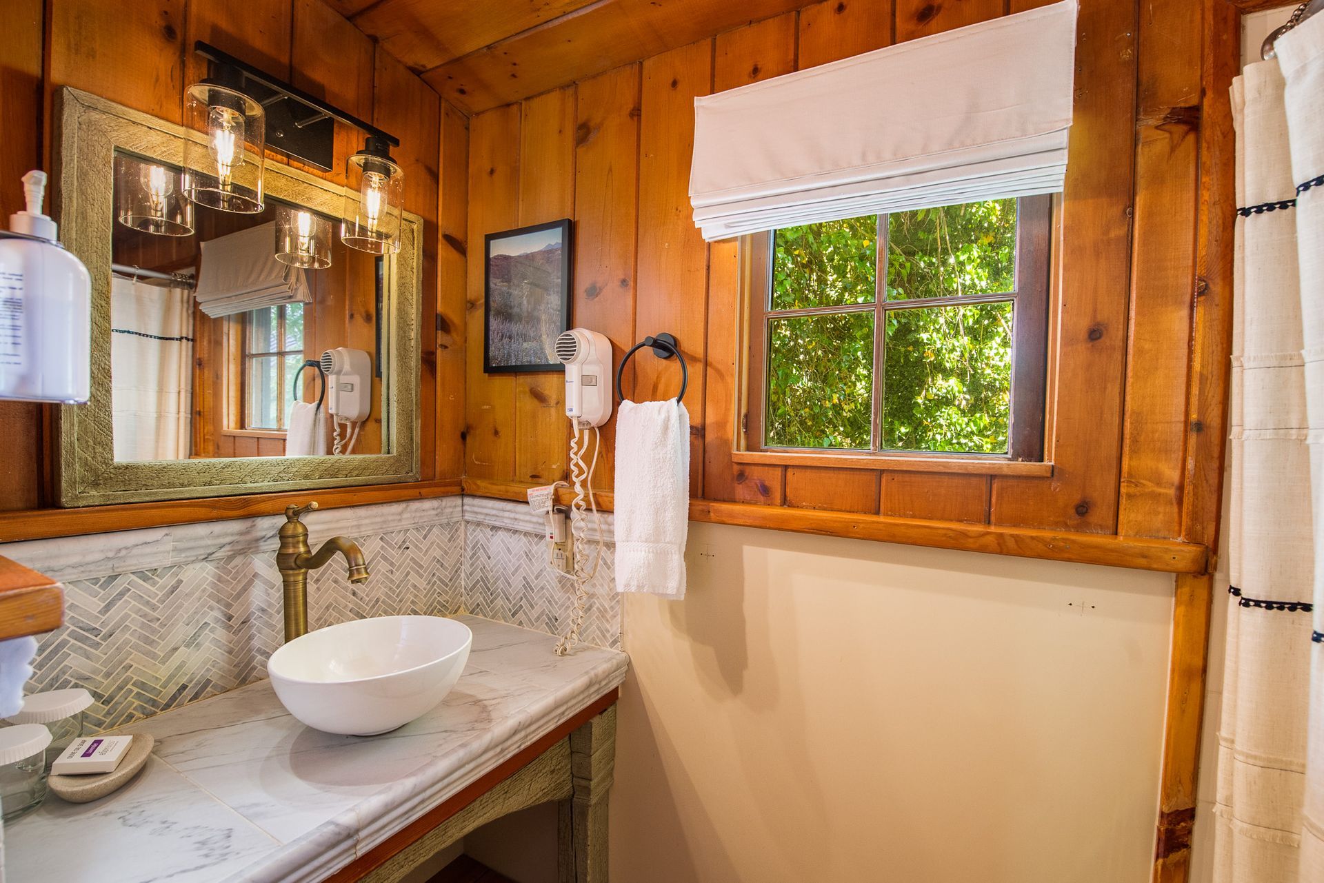 Bathroom with wood-paneled walls, marble countertop, white sink, window, and a decorative mirror.