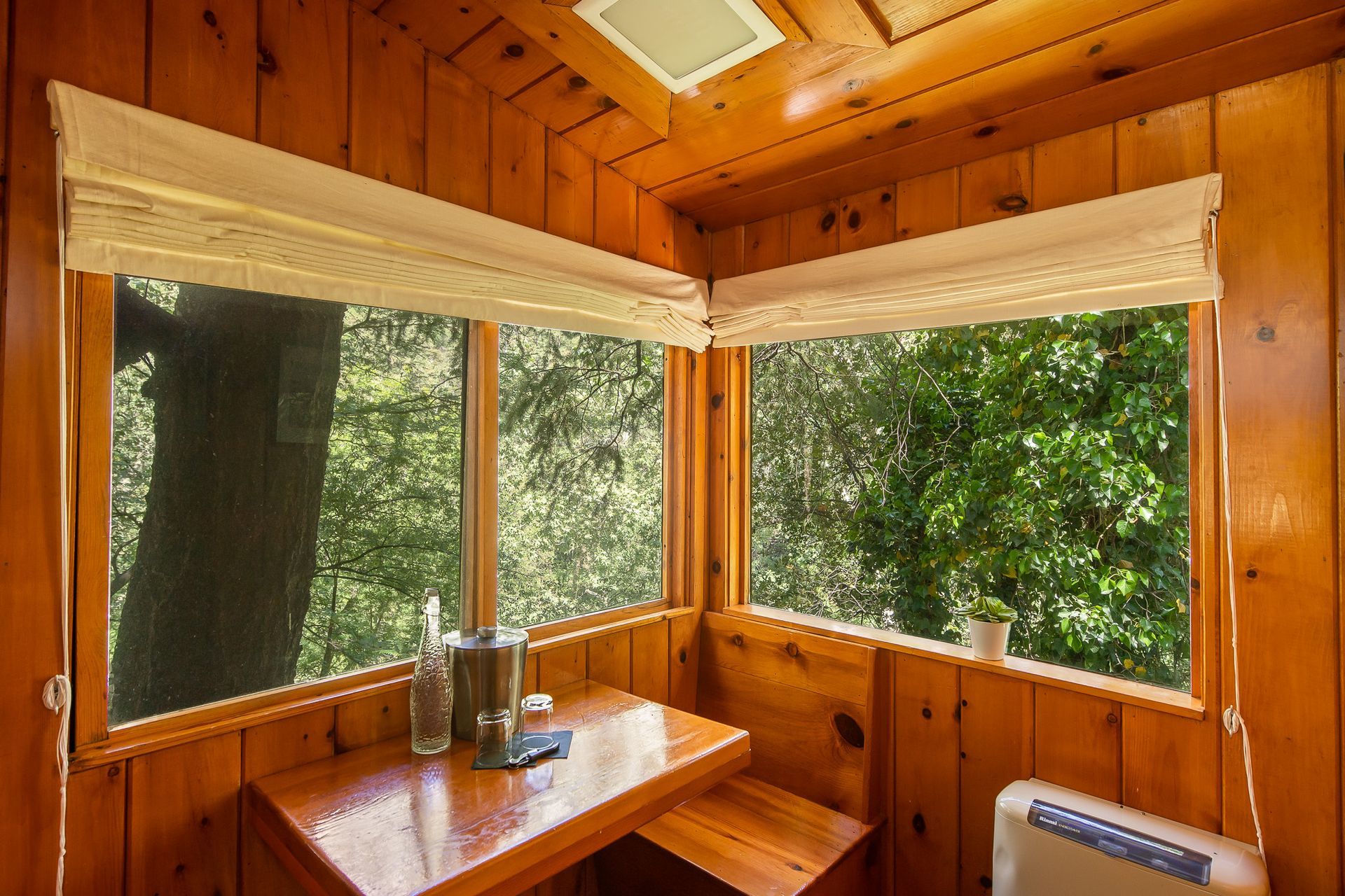 Wooden cabin interior with corner windows overlooking trees, a table with items, and a small heater.