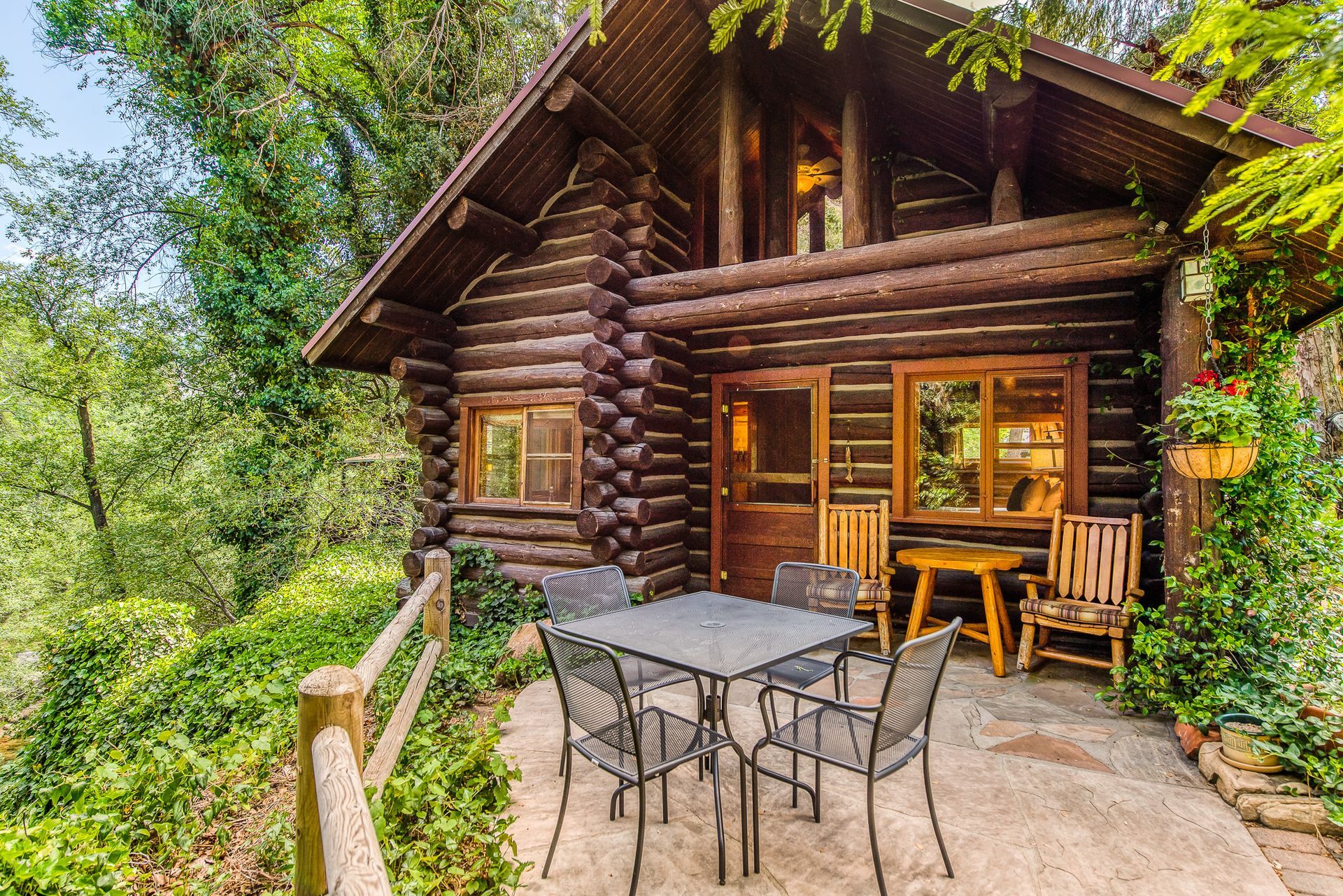 Log cabin with patio, table, chairs, surrounded by trees and foliage.