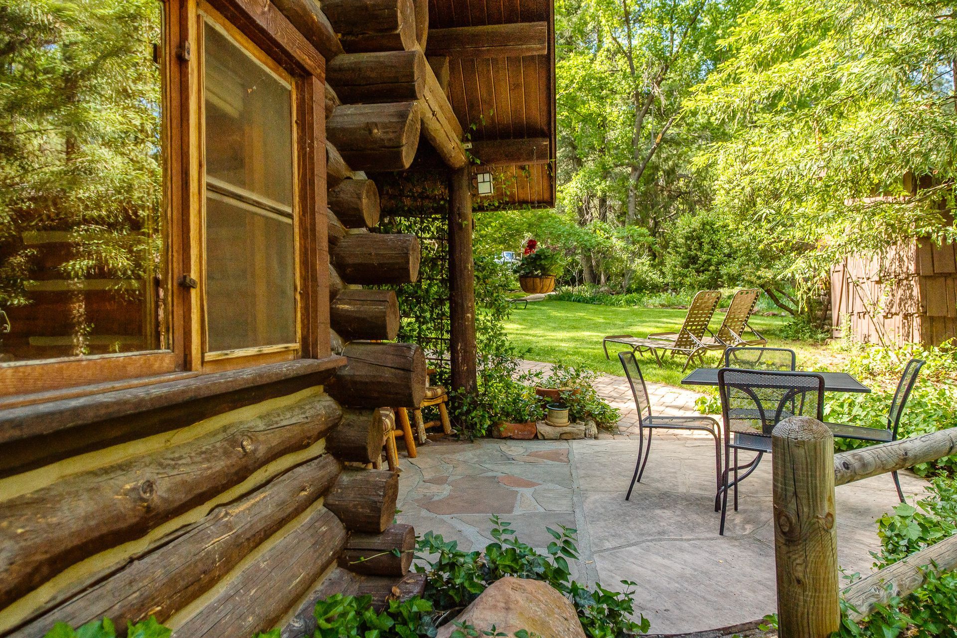 Log cabin exterior with patio, table and chairs, and green lawn in a sunny setting.