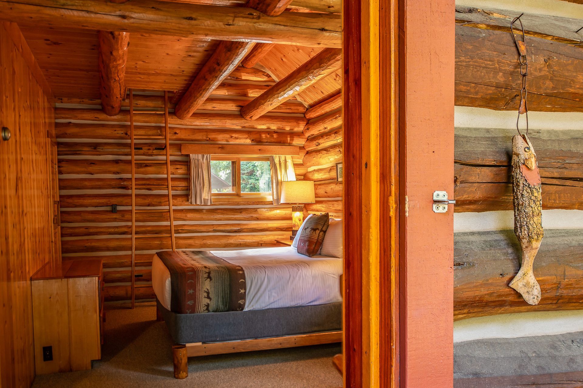Bedroom in a log cabin, seen through a doorway. Bed with patterned blanket, small window, rustic decor.