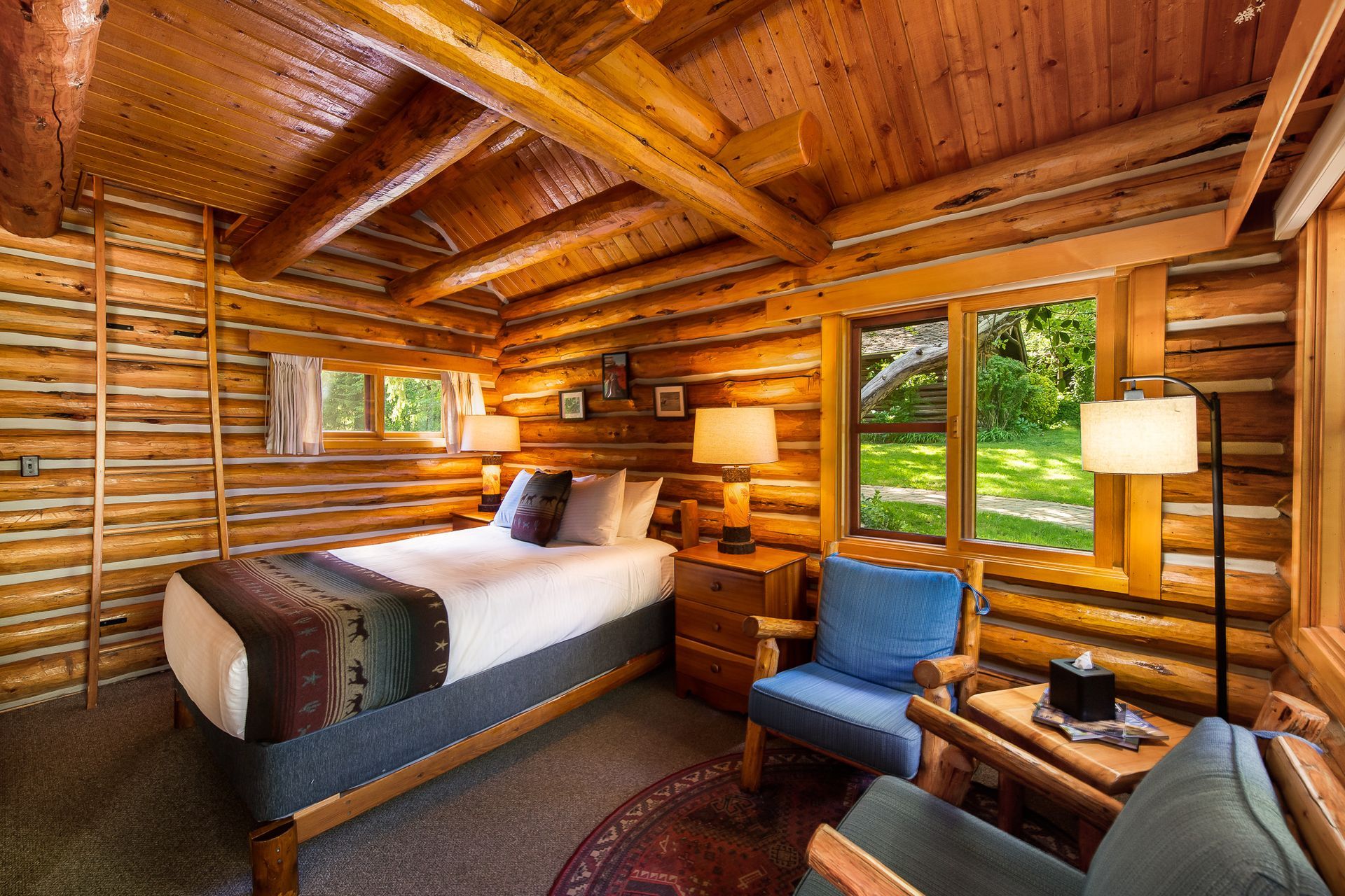 Cozy cabin bedroom with wood-paneled walls and ceiling, bed, chairs, lamps, and small windows overlooking green foliage.