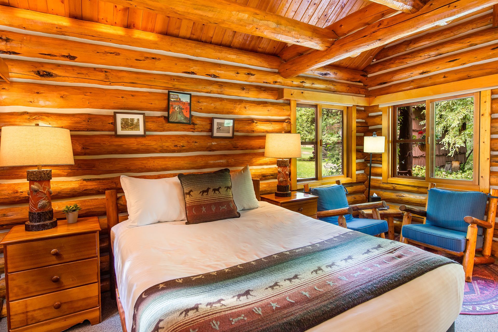 Log cabin bedroom with wood walls, bed, lamps, and blue chairs near windows.
