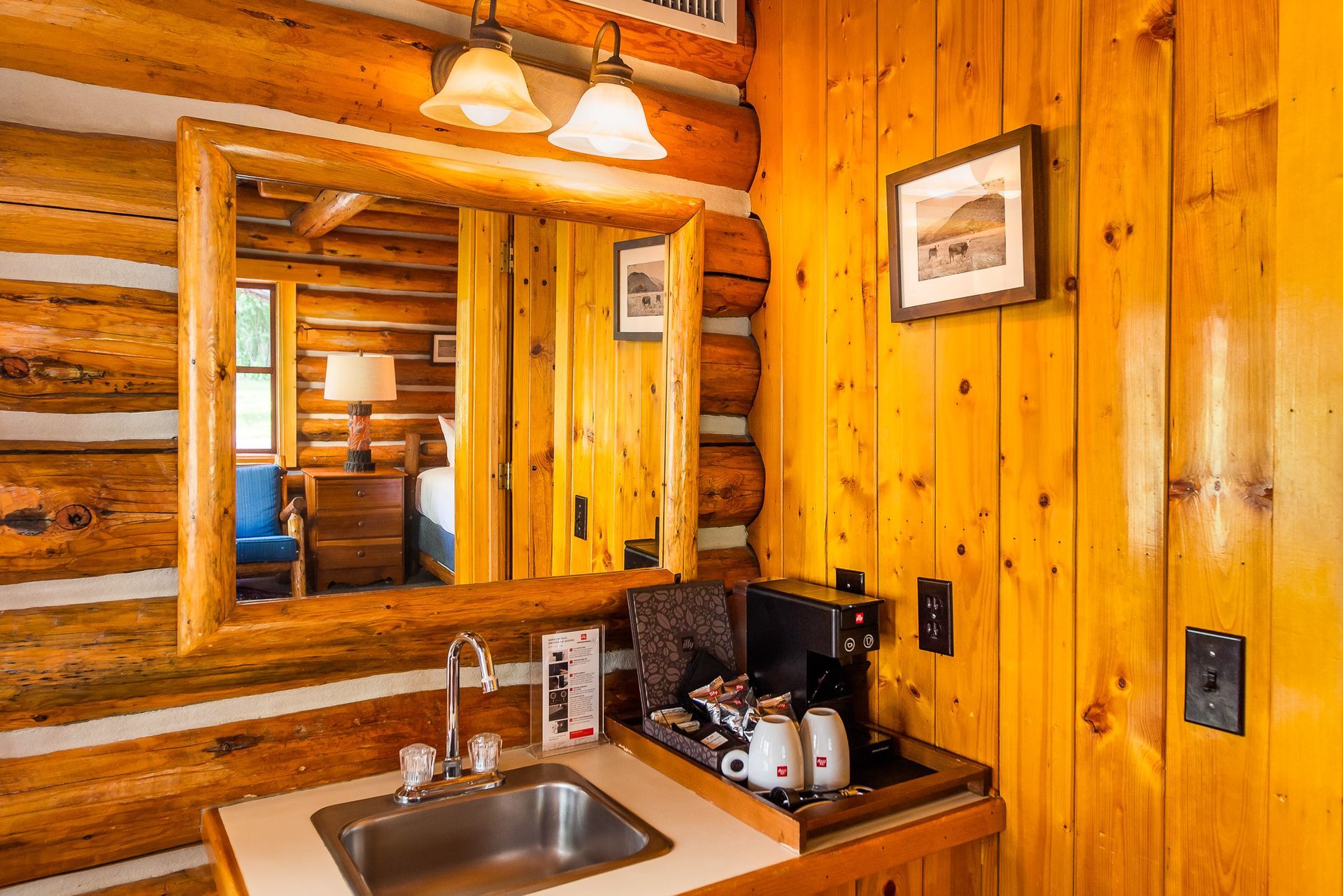 Log-cabin style room interior with sink, mirror, coffee maker, and artwork on wooden walls.
