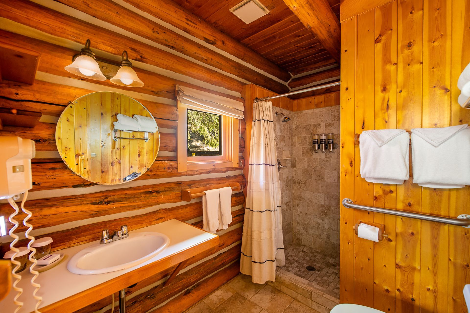 Bathroom with log walls and a shower. White towels, a round mirror, and a window are visible.