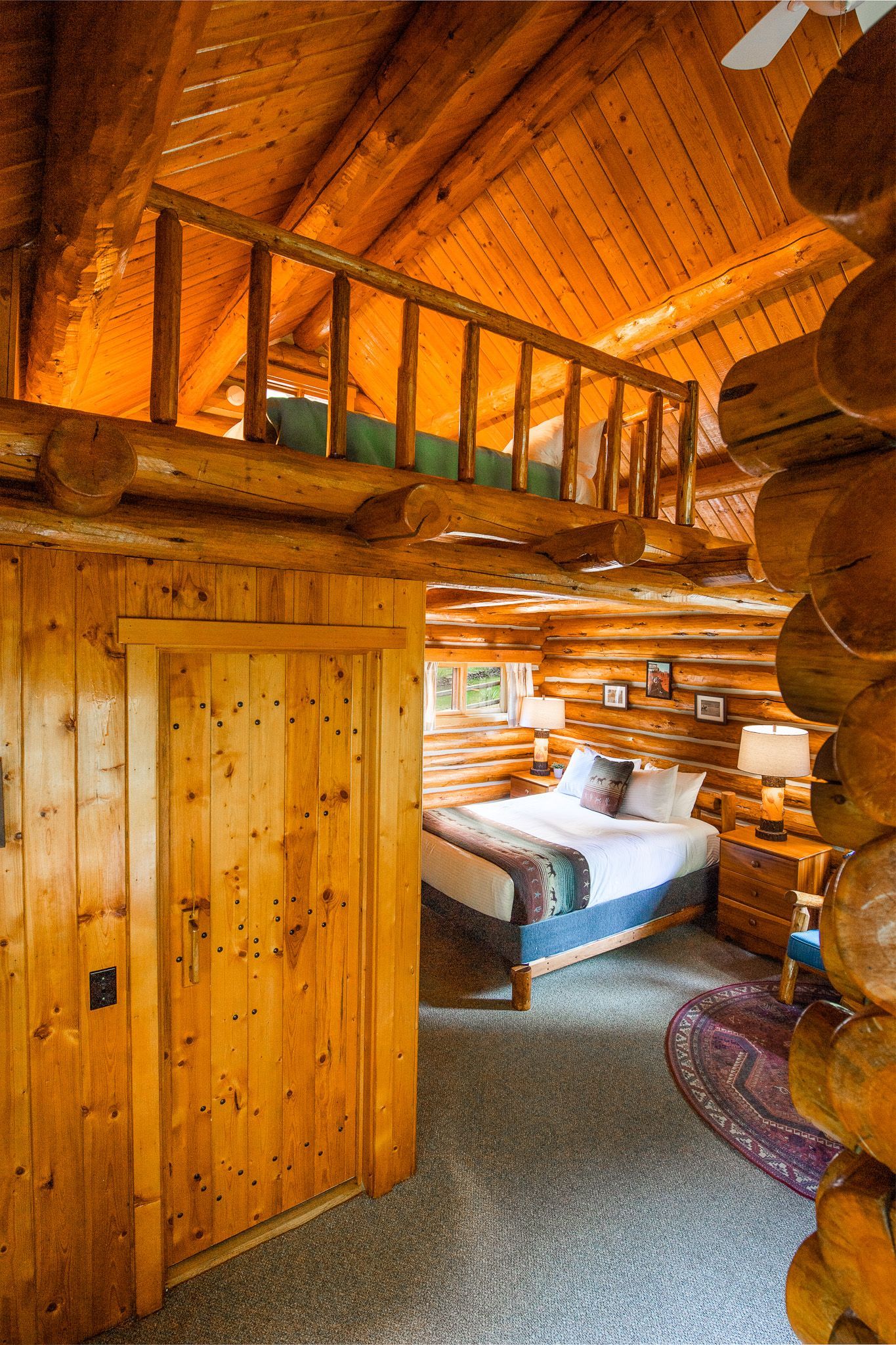 Rustic log cabin interior with a loft, wooden door, and a bed visible.