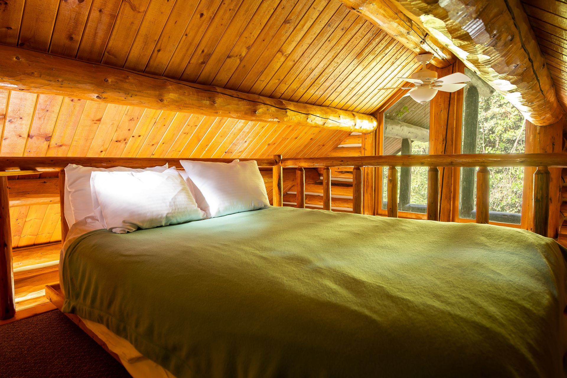 Loft bedroom with log walls, bed, green comforter, and window looking out.