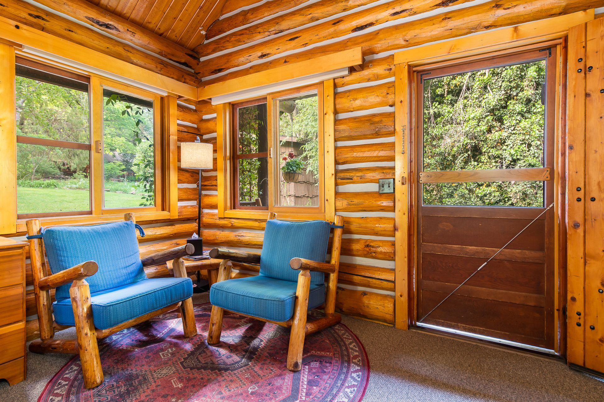 Cozy log cabin interior with blue chairs, a lamp, windows, and a screen door opening to greenery.