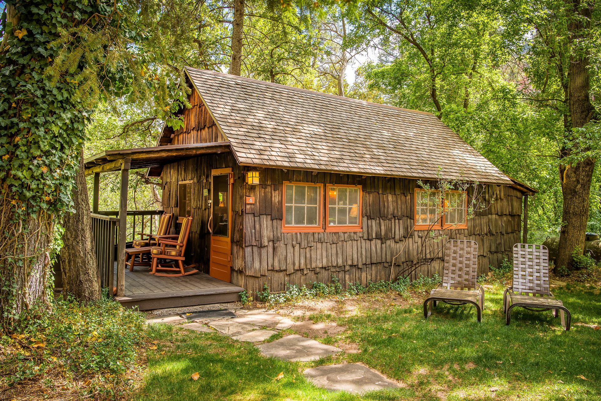 Cozy wooden cabin nestled in a lush green forest, with a porch and rocking chairs.