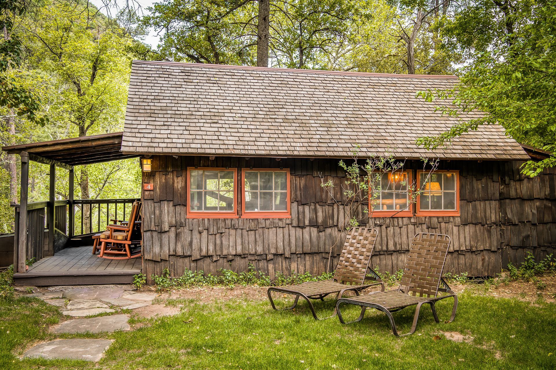 Rustic cabin with porch and two chairs on a grassy lawn, surrounded by trees.