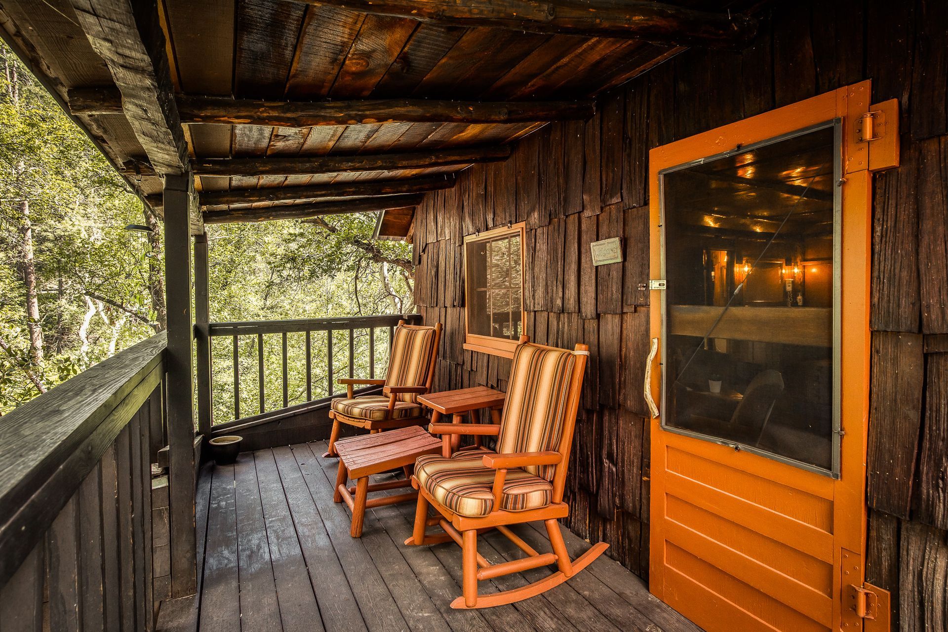 Wooden porch with orange rocking chairs, door, and railing. Trees in background.