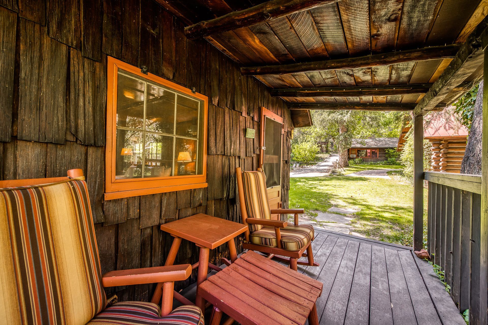 Wooden cabin porch with rocking chairs, orange-framed window, and distant view of yard and other cabins.