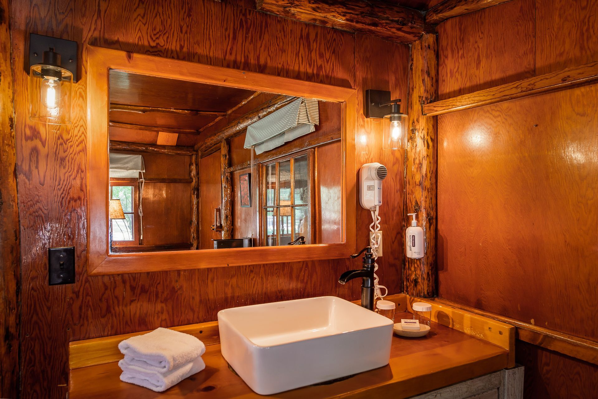 Rustic bathroom with a square sink, wooden paneling, and mirror. Two light fixtures flank the mirror.
