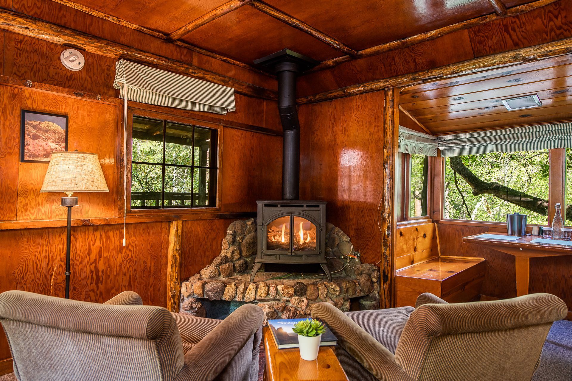 Cozy cabin interior with a wood-burning stove, two armchairs, and a window with a view of trees.
