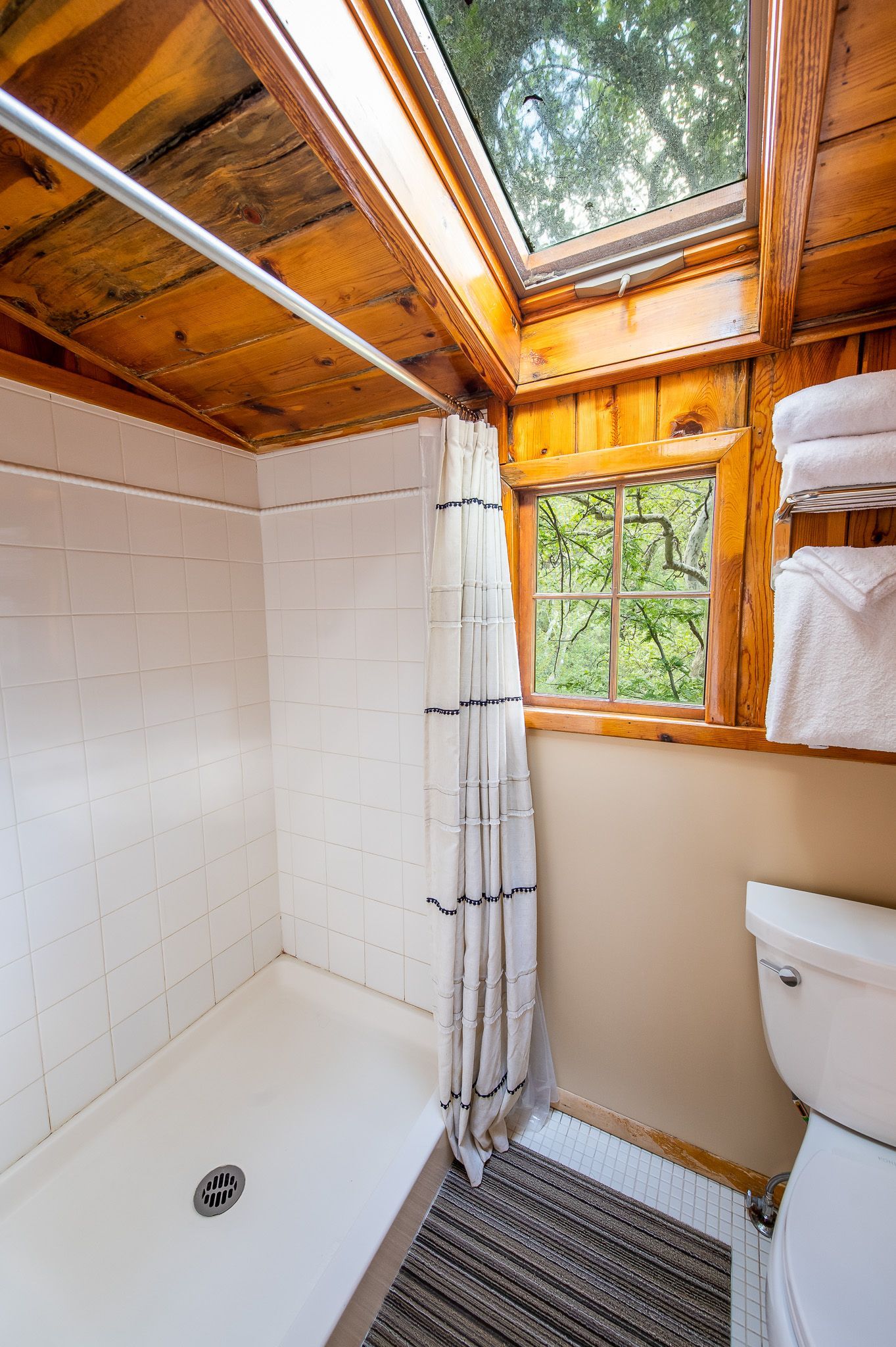 Small cabin bathroom with shower, toilet, window, and wooden ceiling and trim.