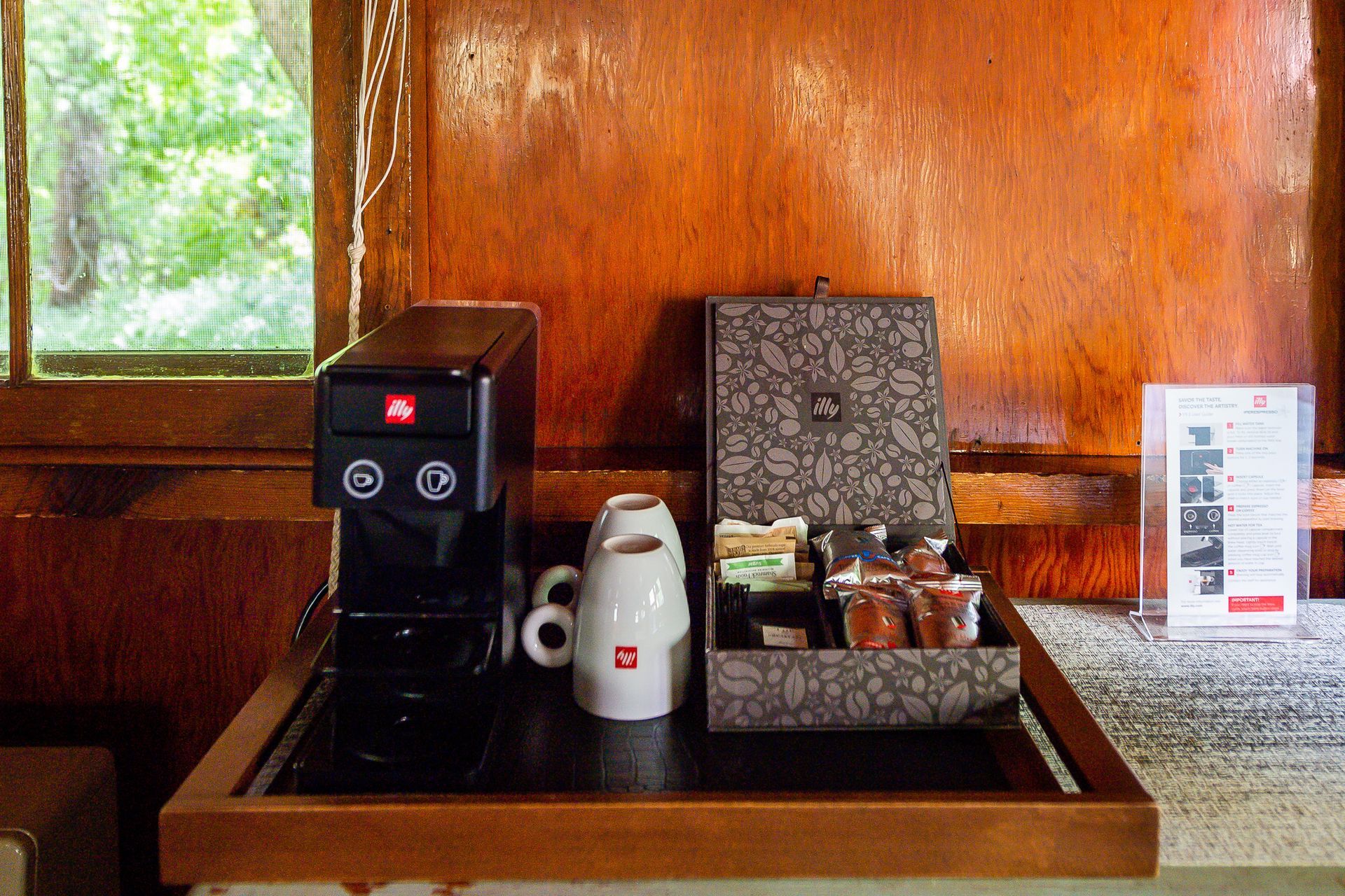 Coffee station with a black espresso machine, cup, and a box of coffee supplies on a wooden tray.