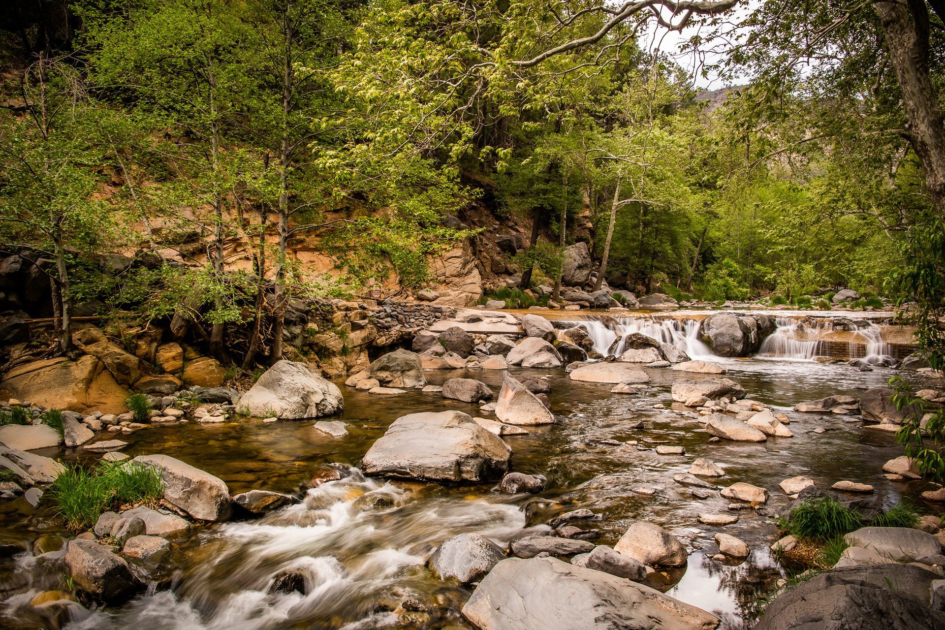 A shallow river flows over rocks, with a small waterfall, surrounded by green trees in a natural setting.