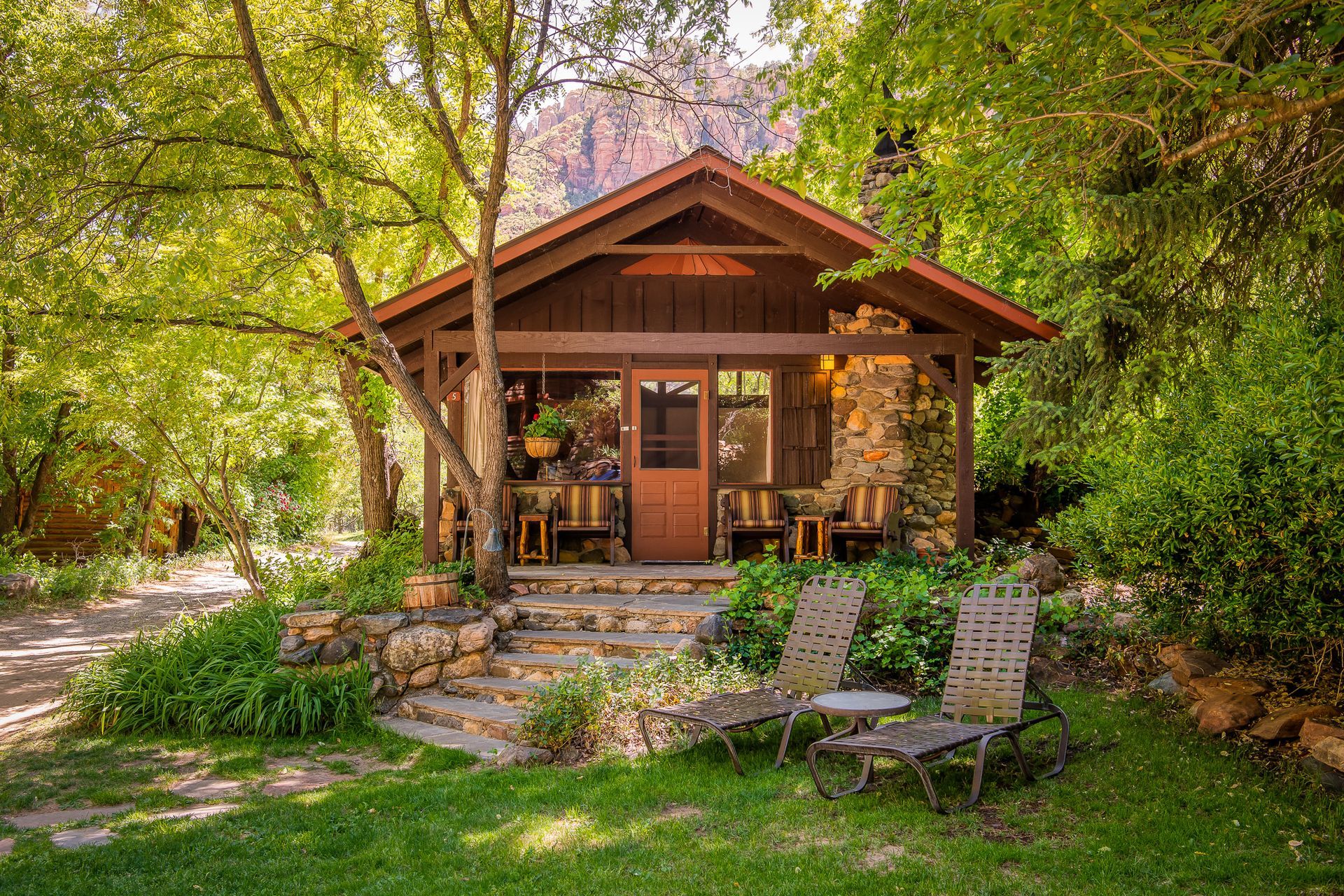 Cozy cabin with stone chimney, porch, and chairs on the lawn, surrounded by trees.