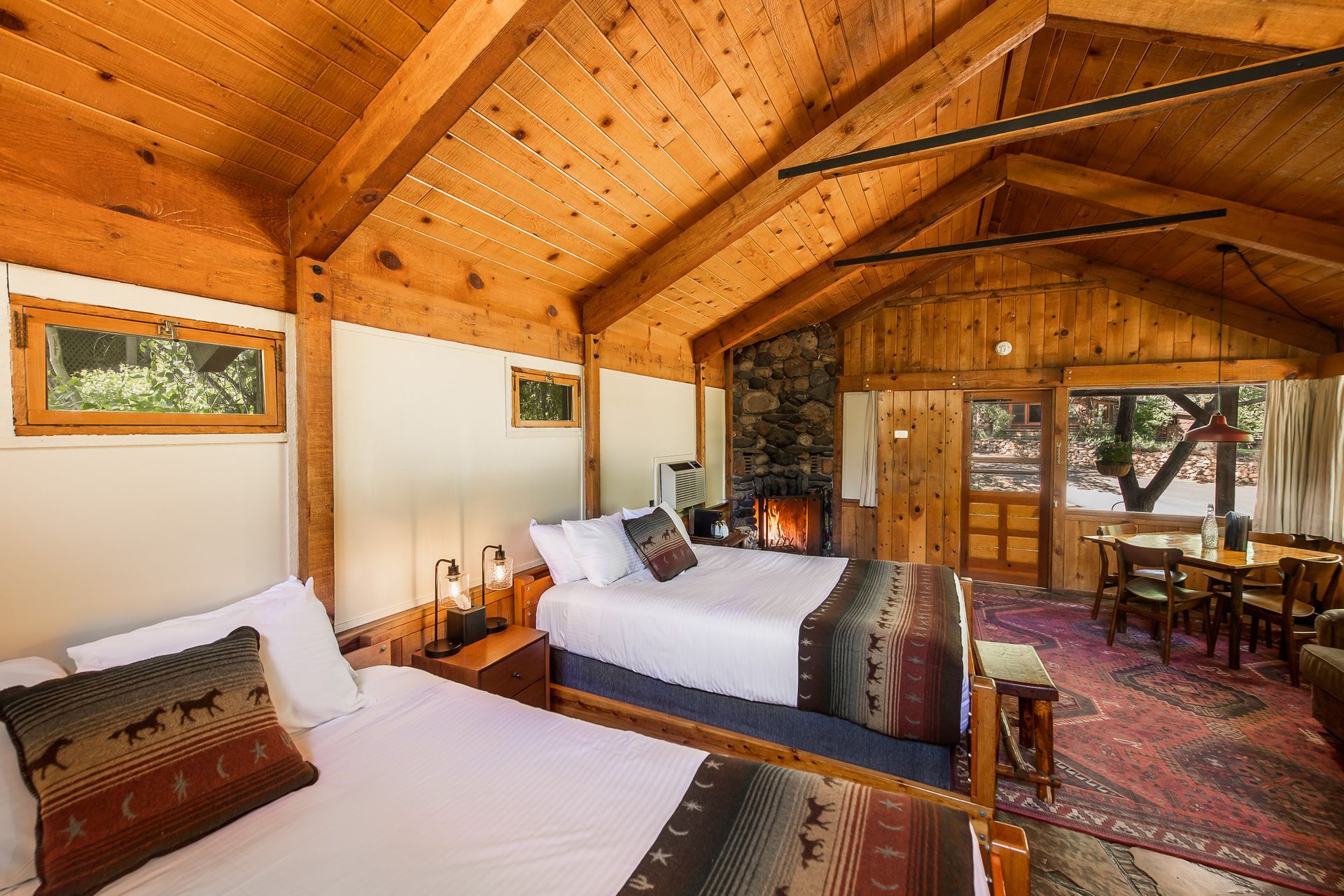 Rustic cabin interior with two beds, fireplace, dining area, and wood beams.