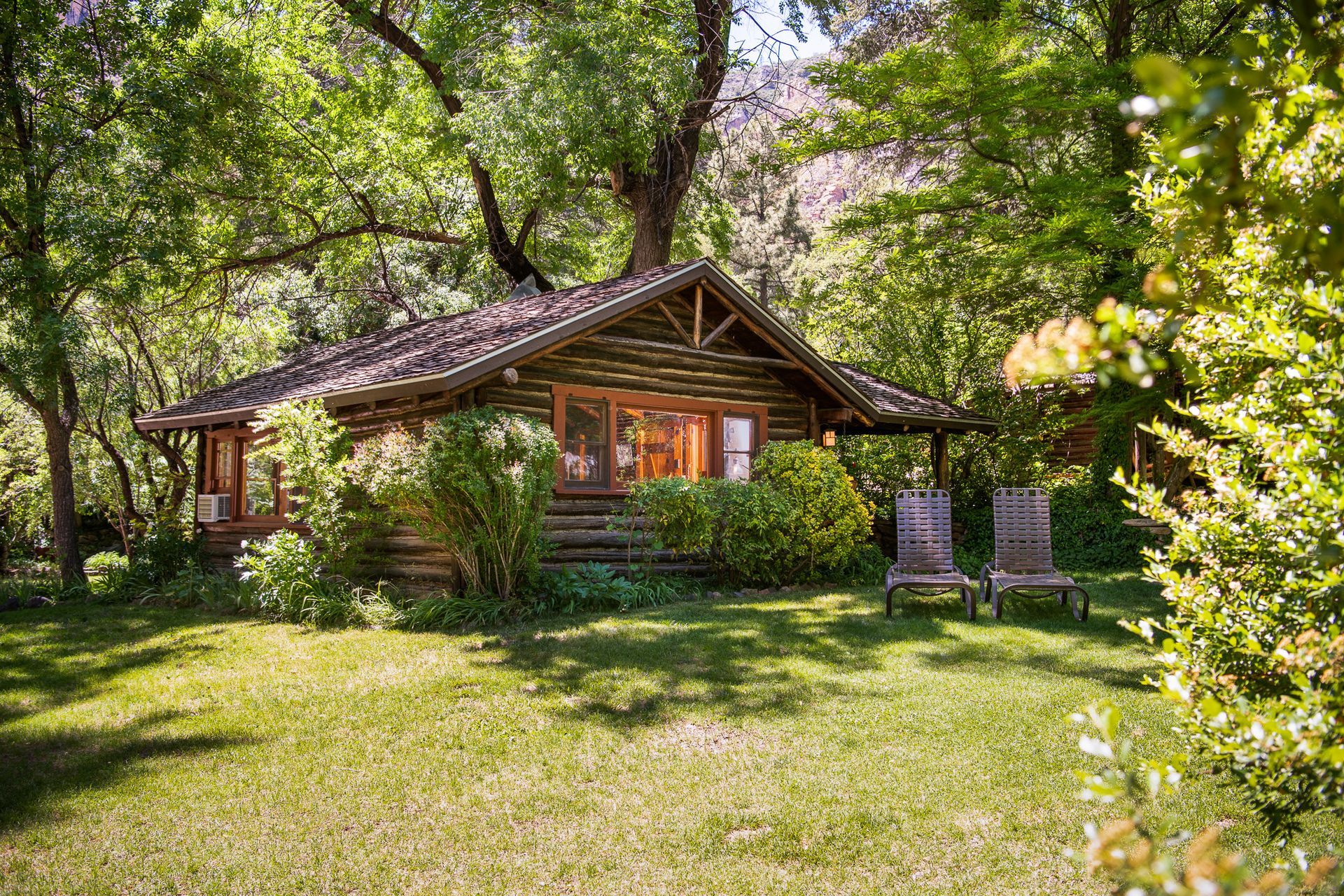 Log cabin nestled in a green, leafy setting with two chairs on the grass.