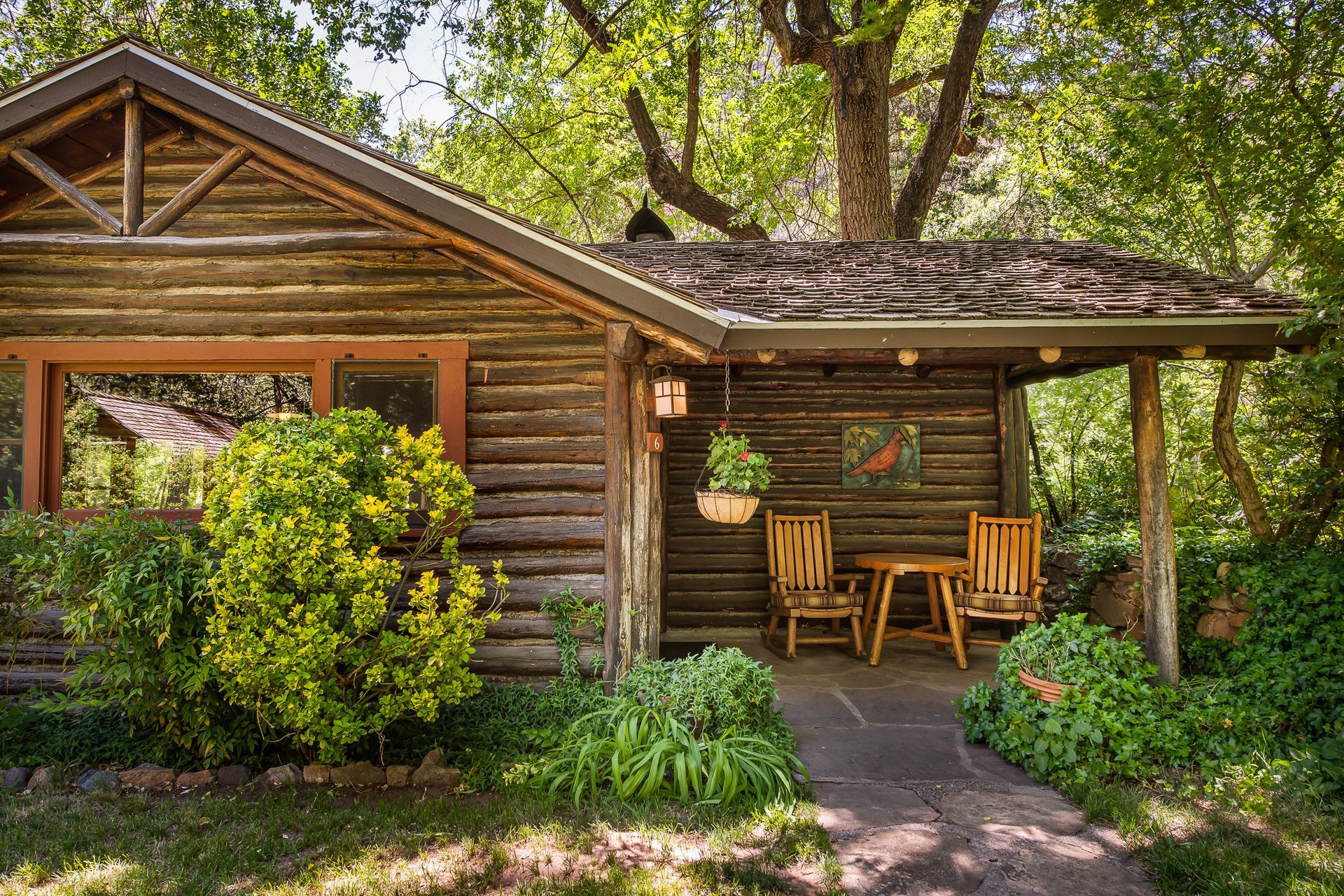 Wooden cabin with porch, table, chairs, surrounded by greenery.