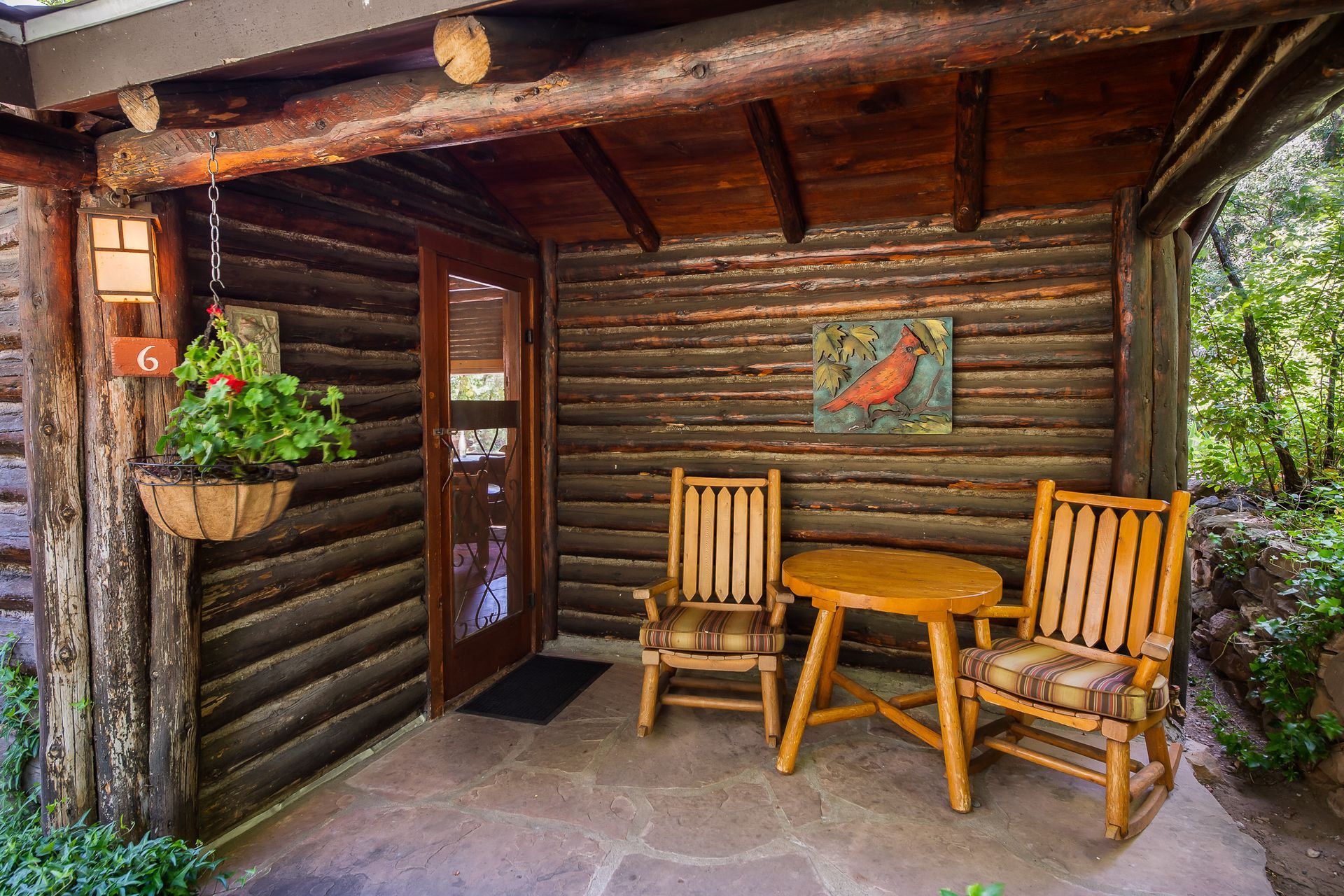 Log cabin porch with wooden chairs and small table; red bird art and hanging basket.