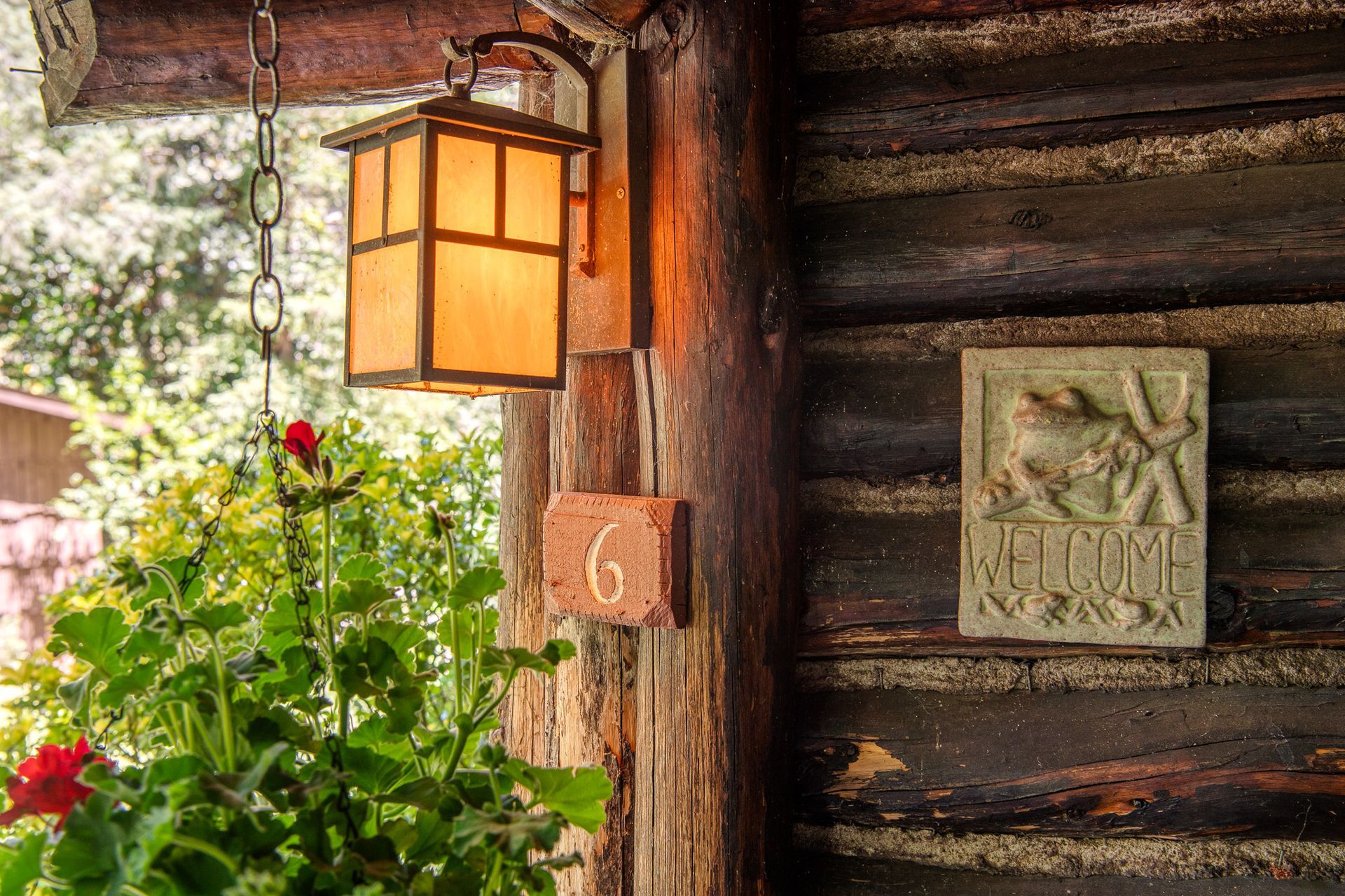 Log cabin entrance with lantern, hanging flower basket, house number 6, and a welcome sign.