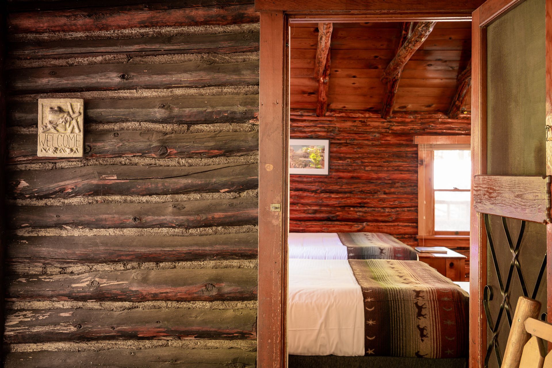 A rustic cabin bedroom interior. Two beds visible through a doorway, log walls, wood ceiling.