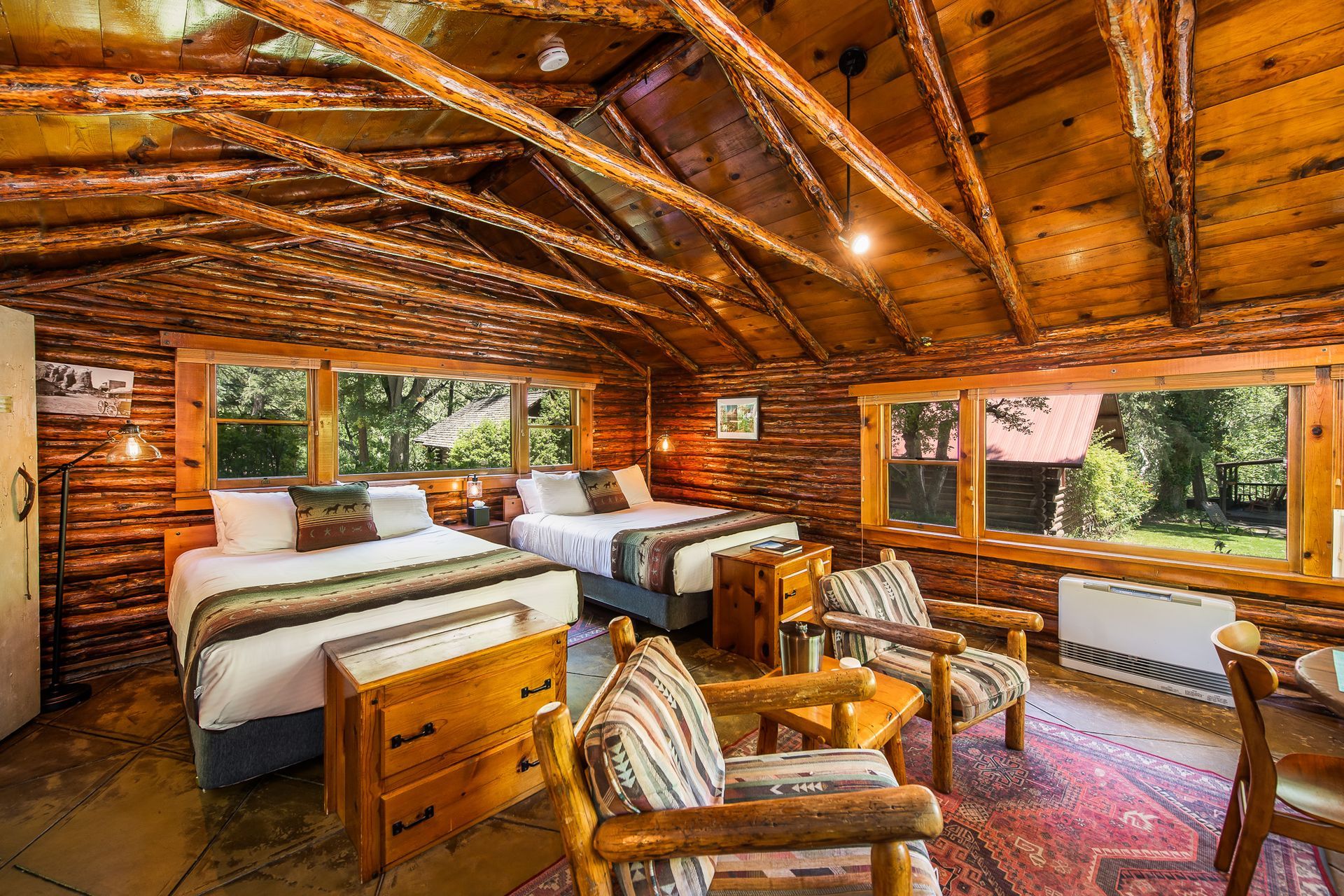 Rustic cabin bedroom with two beds, two chairs, wooden walls, and windows.