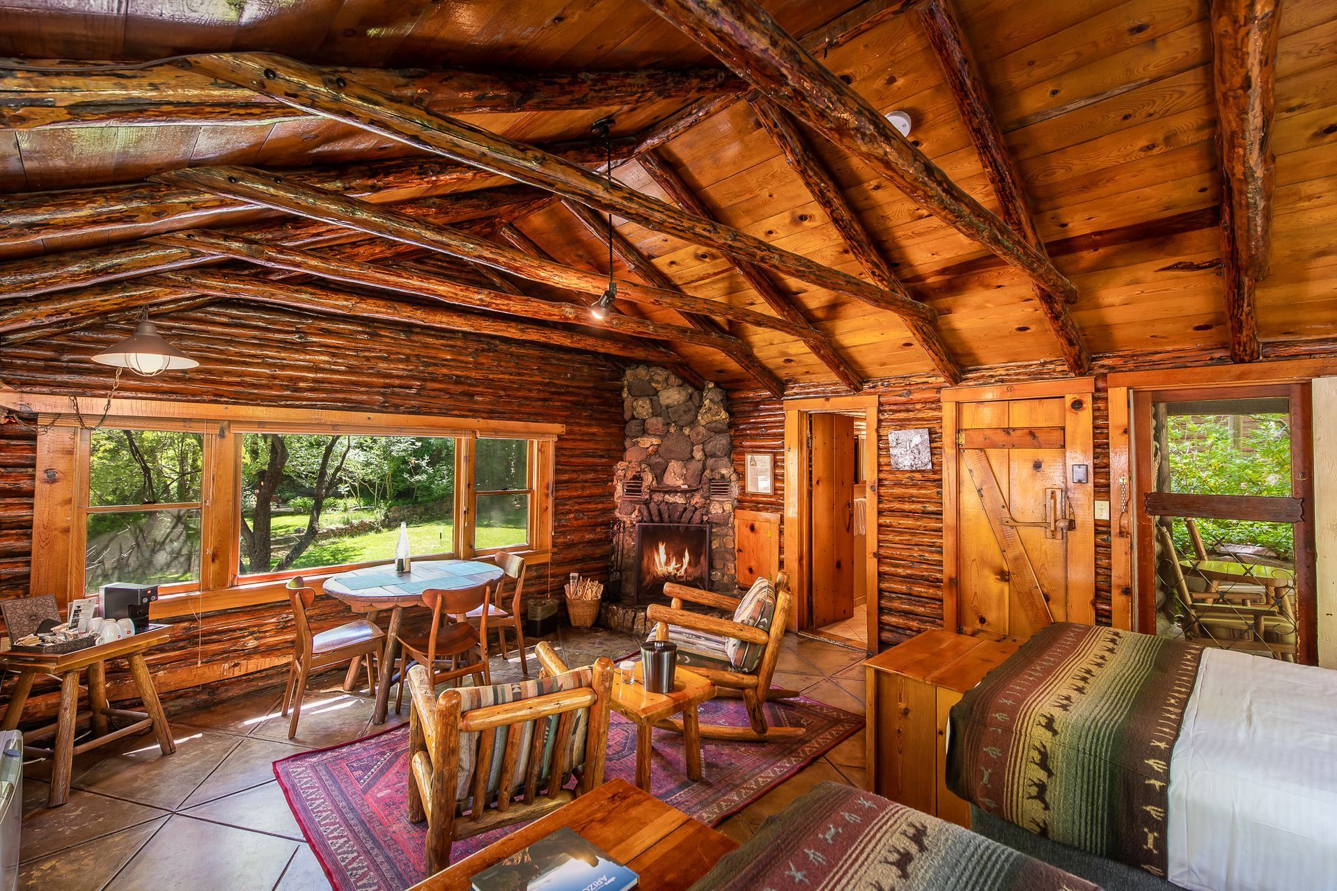 Cozy log cabin interior with wooden furniture, fireplace, and windows overlooking greenery.