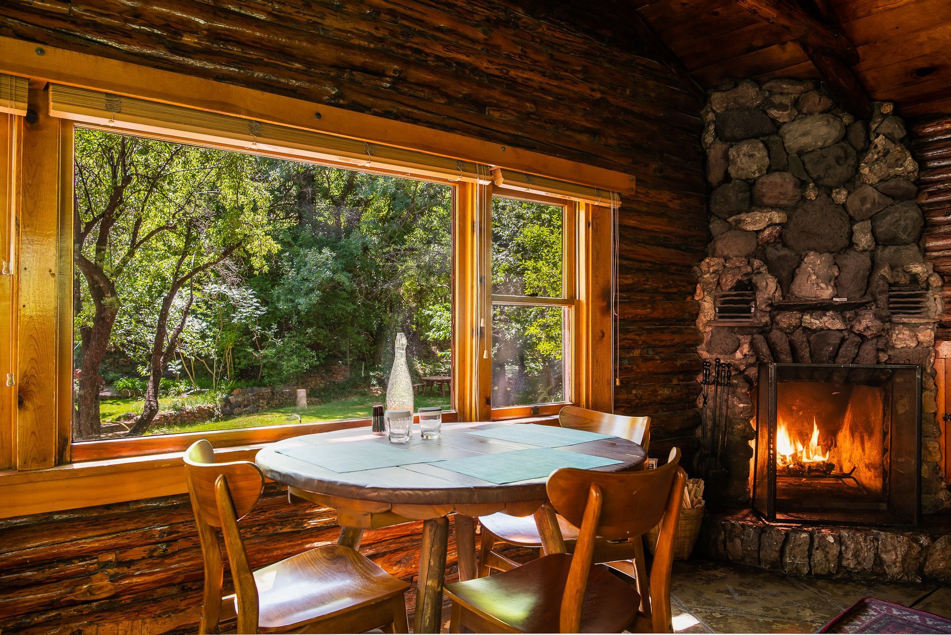 Interior of a cabin with a round table by a window overlooking trees and a lit fireplace.