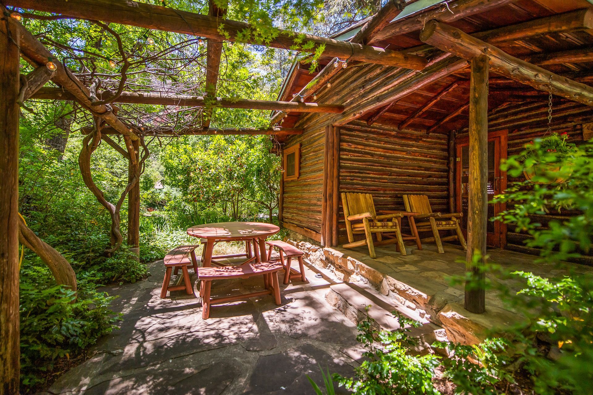 Log cabin with outdoor seating under a wooden pergola, surrounded by lush greenery.