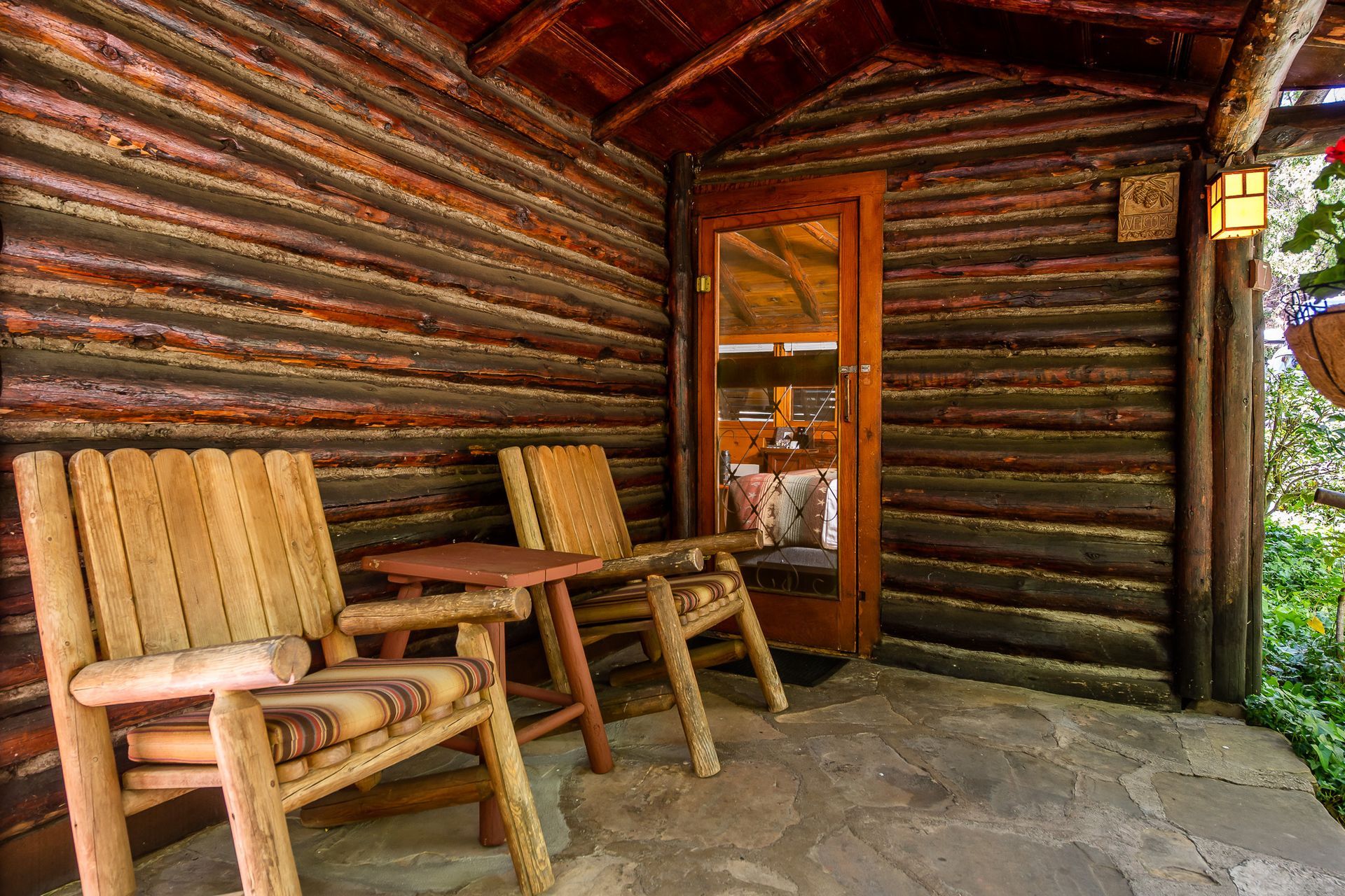 Wooden cabin porch with two chairs, small table, and a door with a reflection.