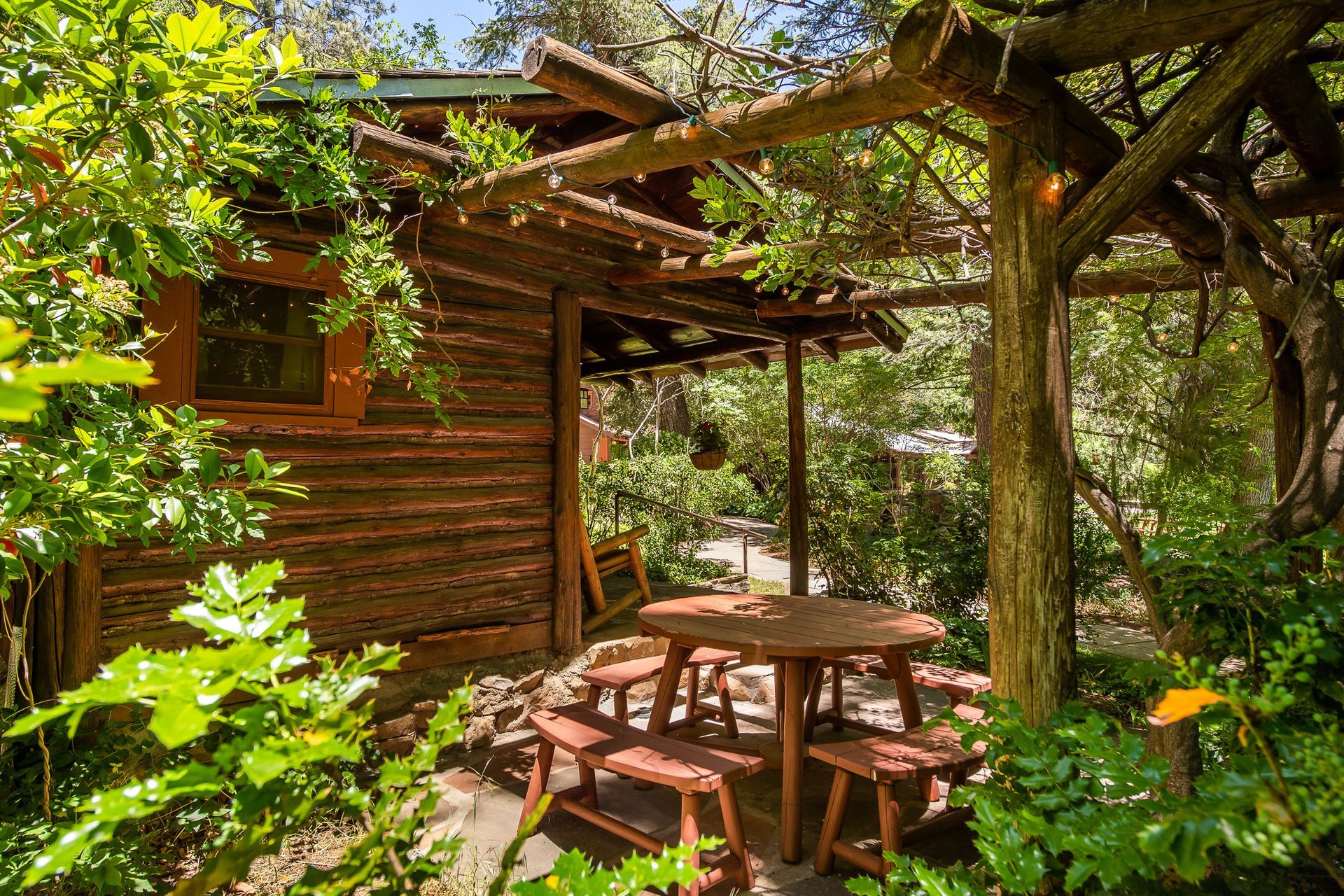 Wooden cabin with a rustic table and benches under a pergola, surrounded by lush greenery.